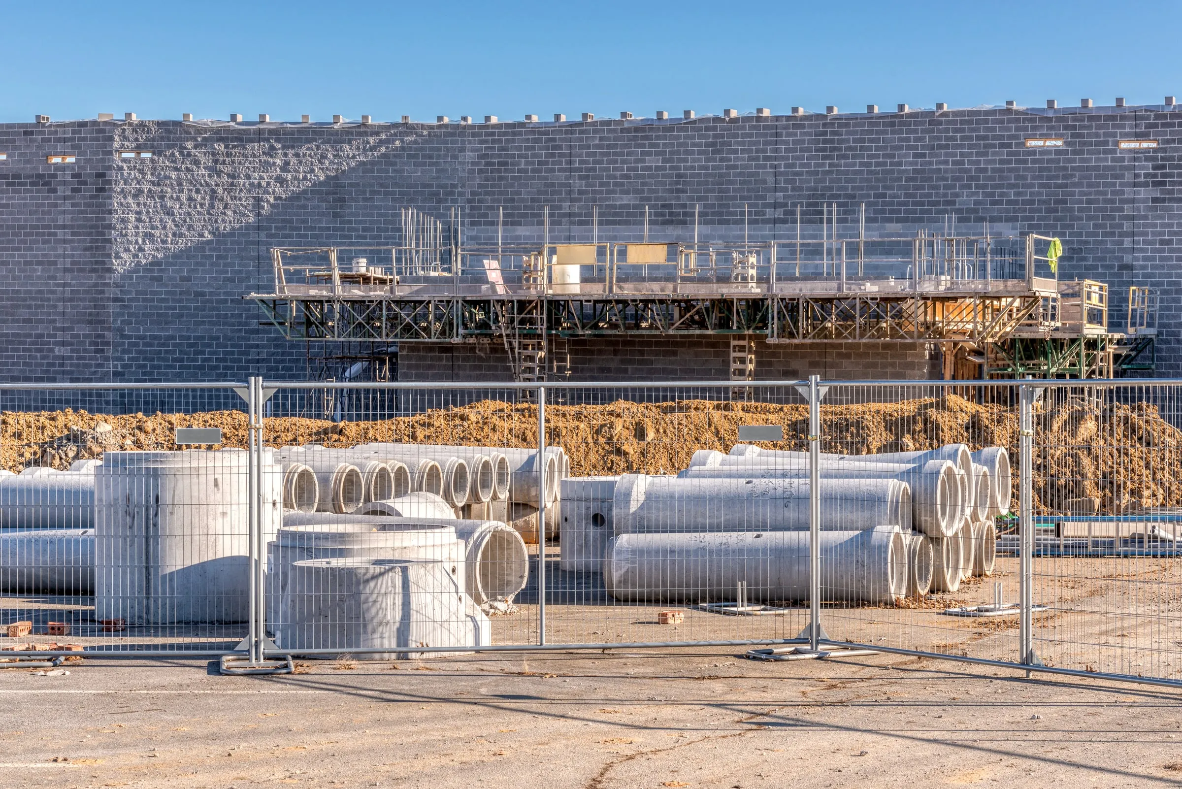 Temporary chain-link fencing around stacked precast concrete pipe sections at a job site, with a block building and a scaffold platform behind it