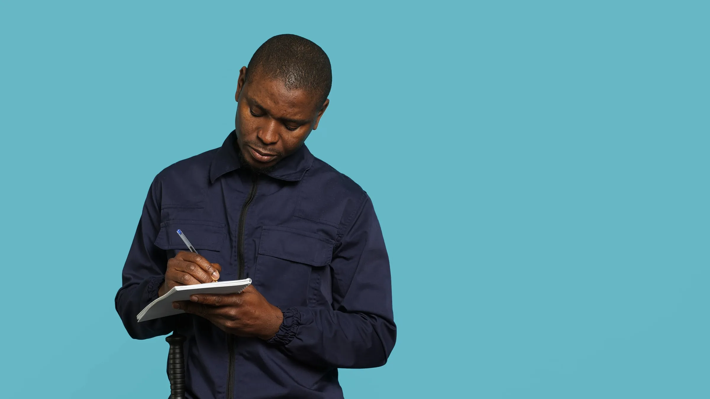 Man in a dark uniform jacket writing in a small spiral notebook against a plain blue background