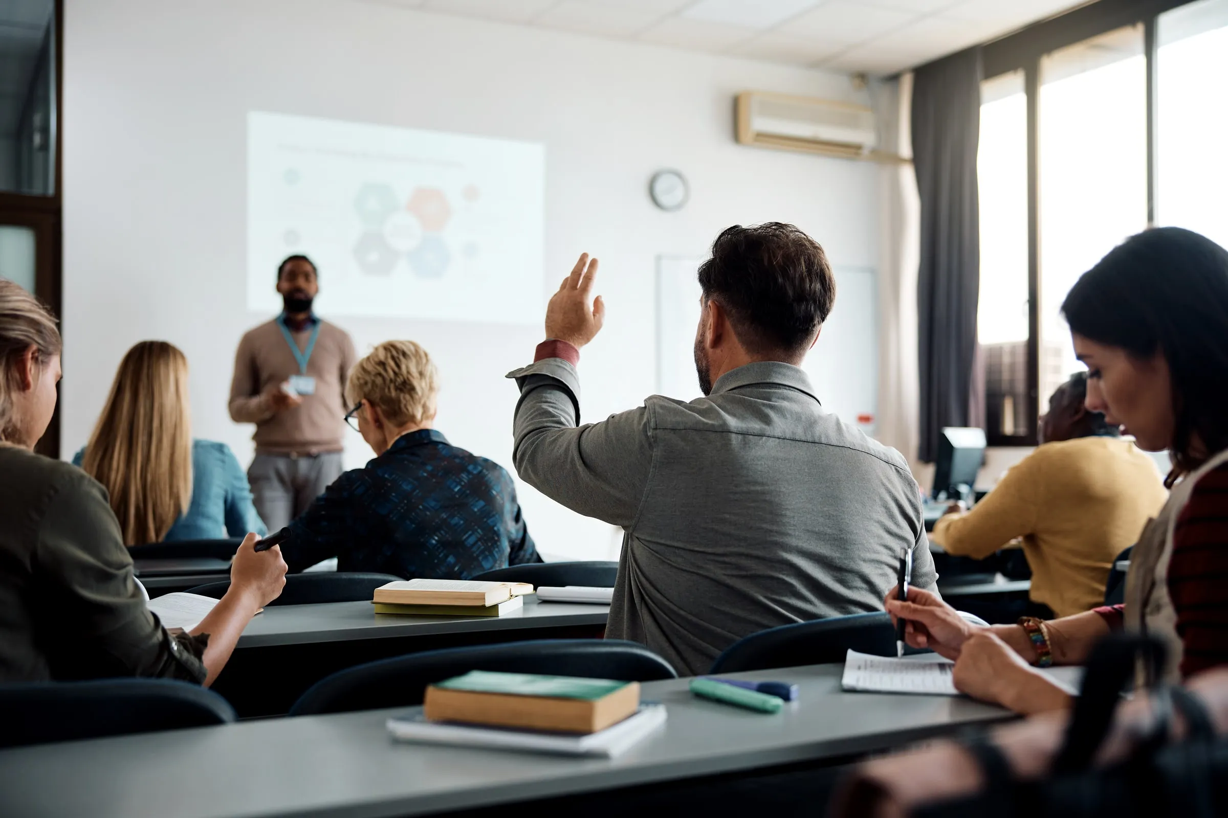 Man raising a hand to ask a question in a classroom while an instructor gestures beside a screen showing a hexagon diagram
