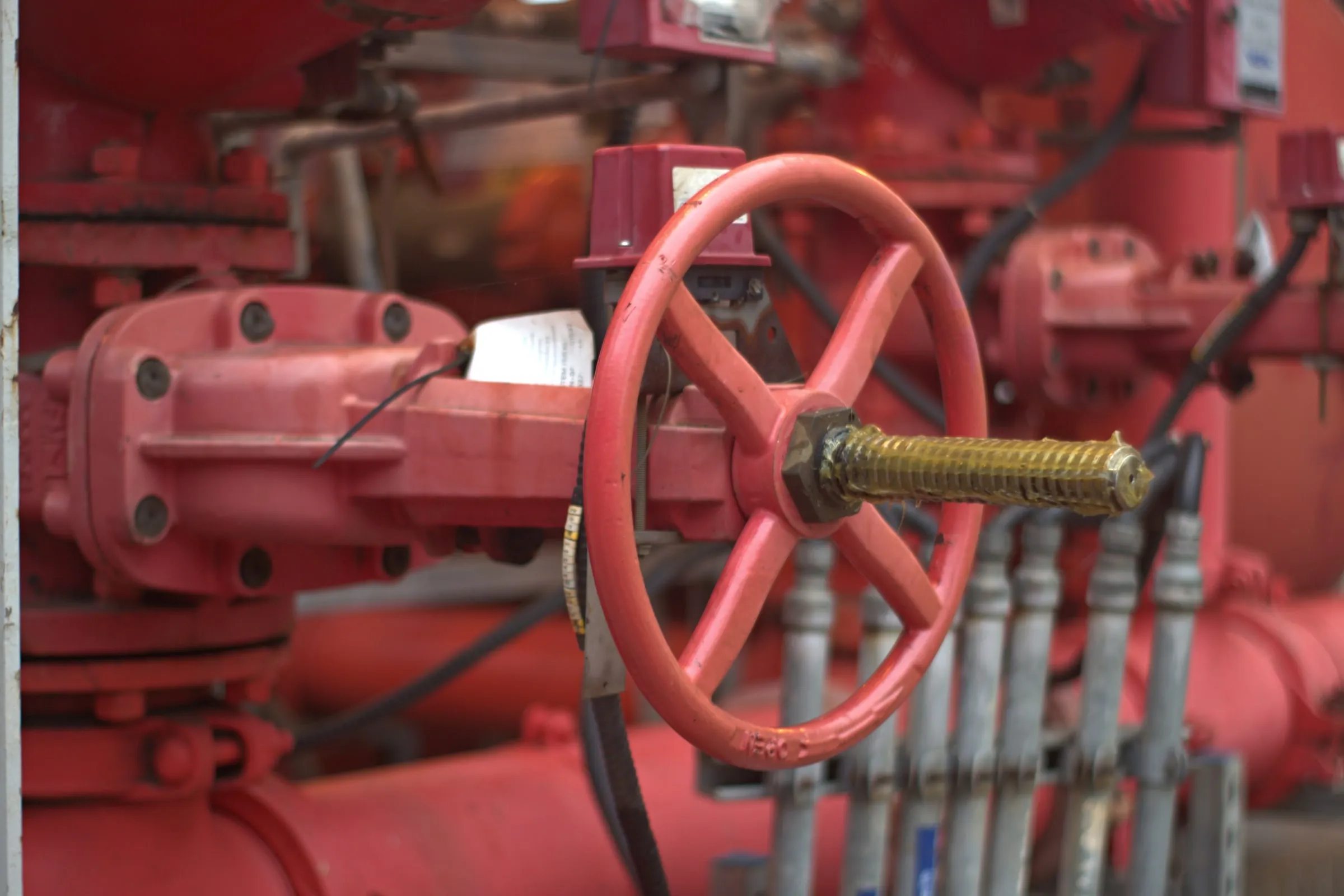 Close-up of a red fire pump valve wheel and brass stem among red risers, gauges and hose connections in a mechanical room