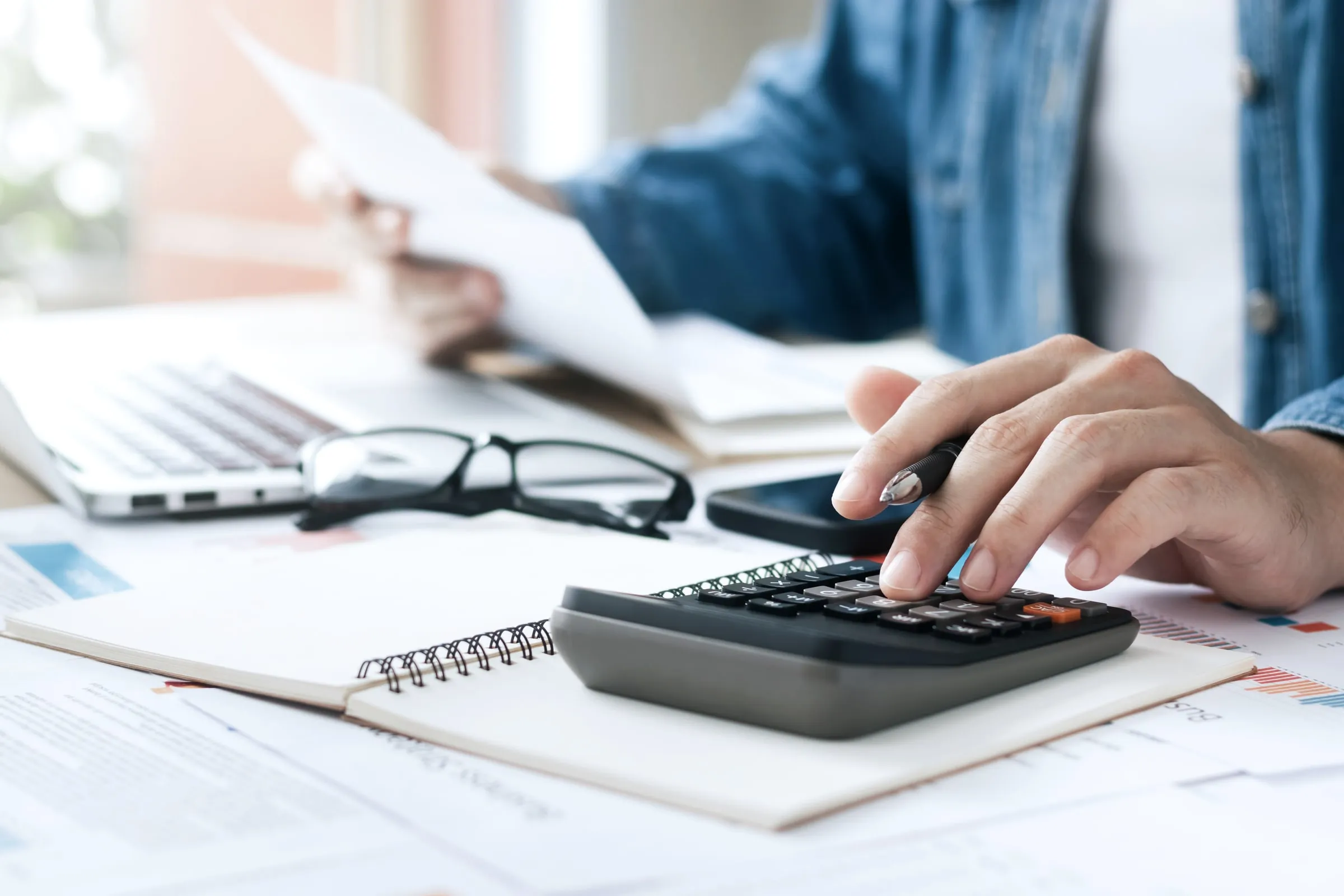 Person in a denim shirt uses a calculator beside a notebook, glasses and a laptop while reviewing paperwork at a desk
