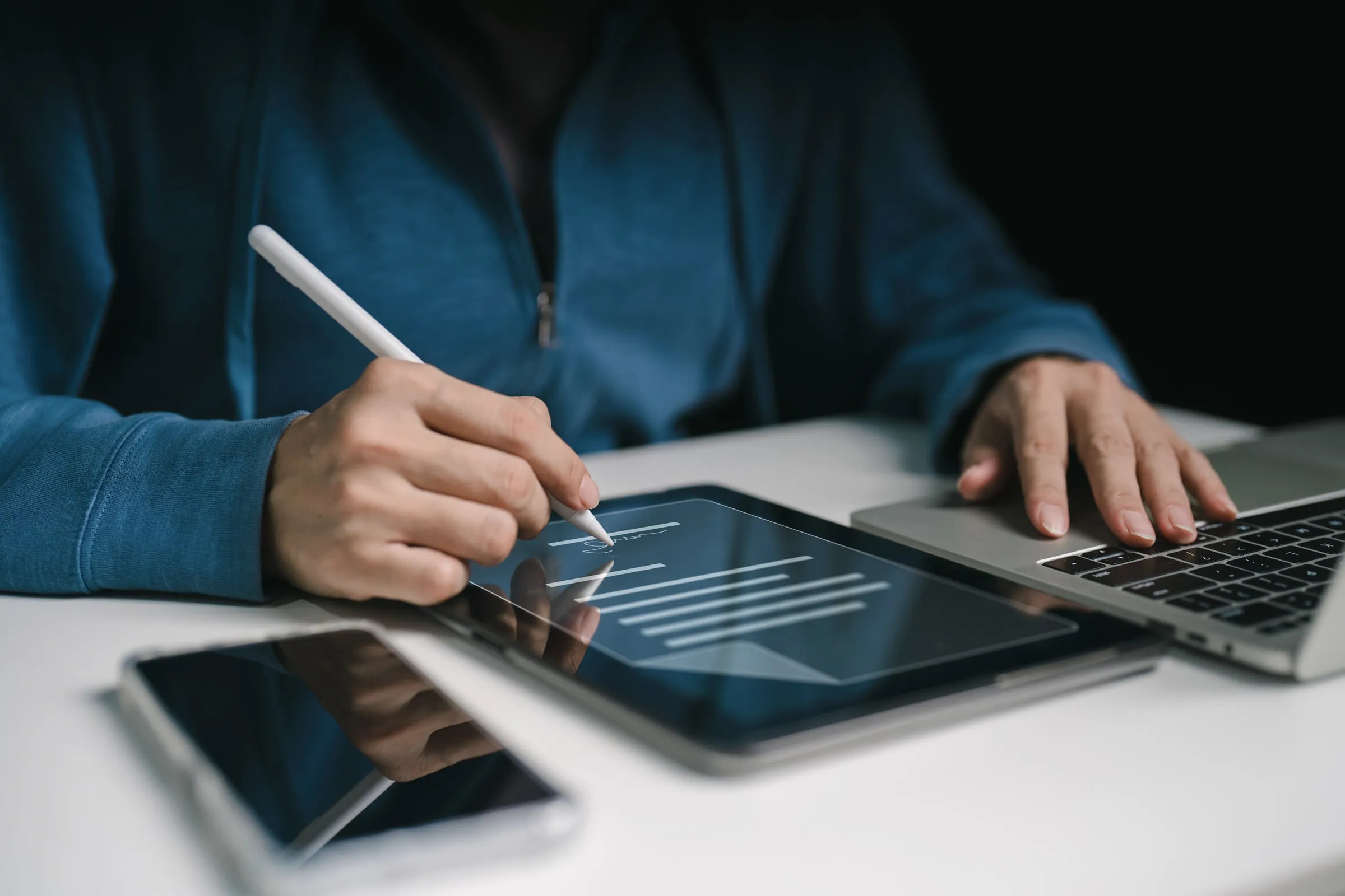 Person using a stylus to sign a document on a tablet screen next to an open laptop and a smartphone