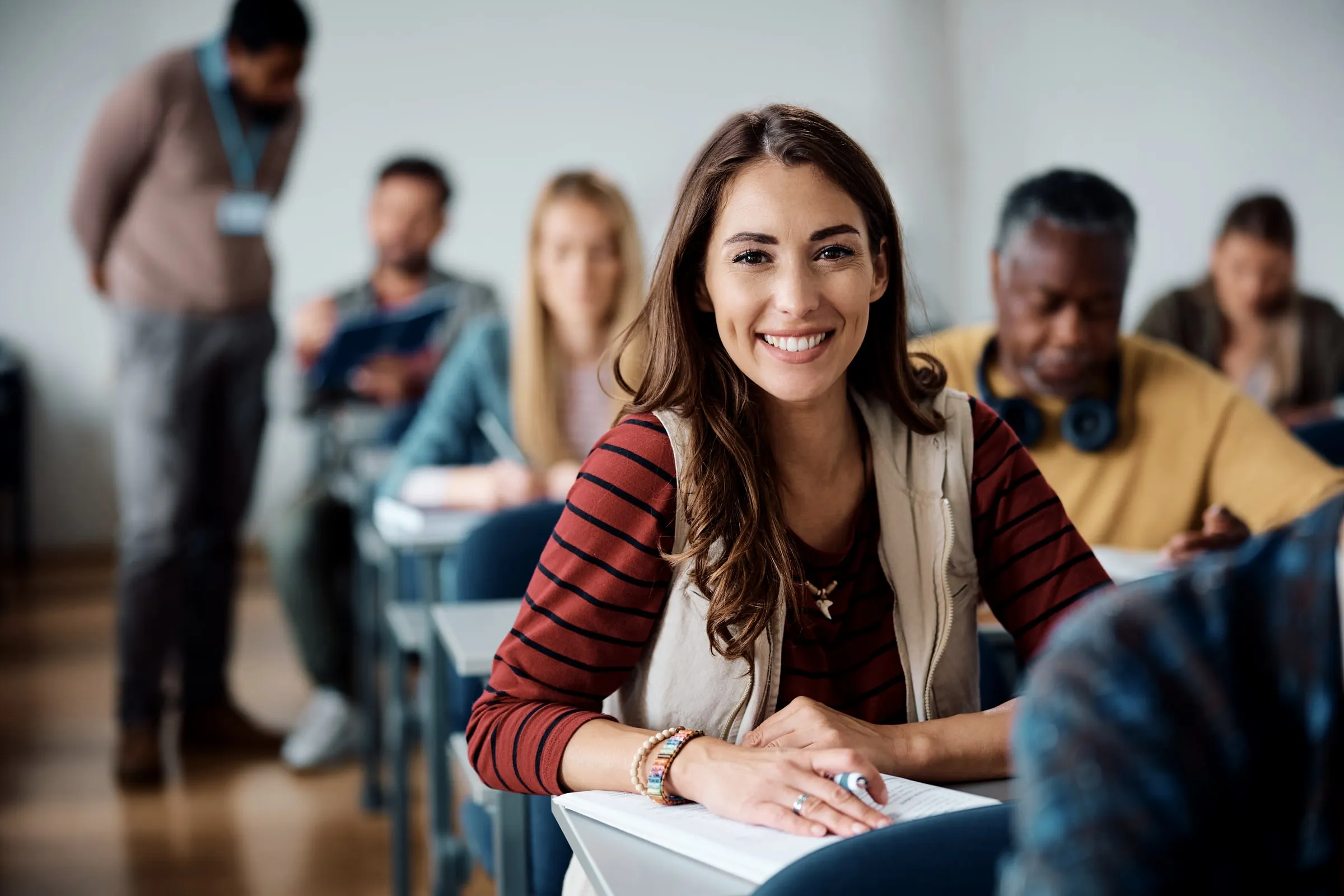 Smiling woman at a classroom desk with other adult students and an instructor with a lanyard in the background