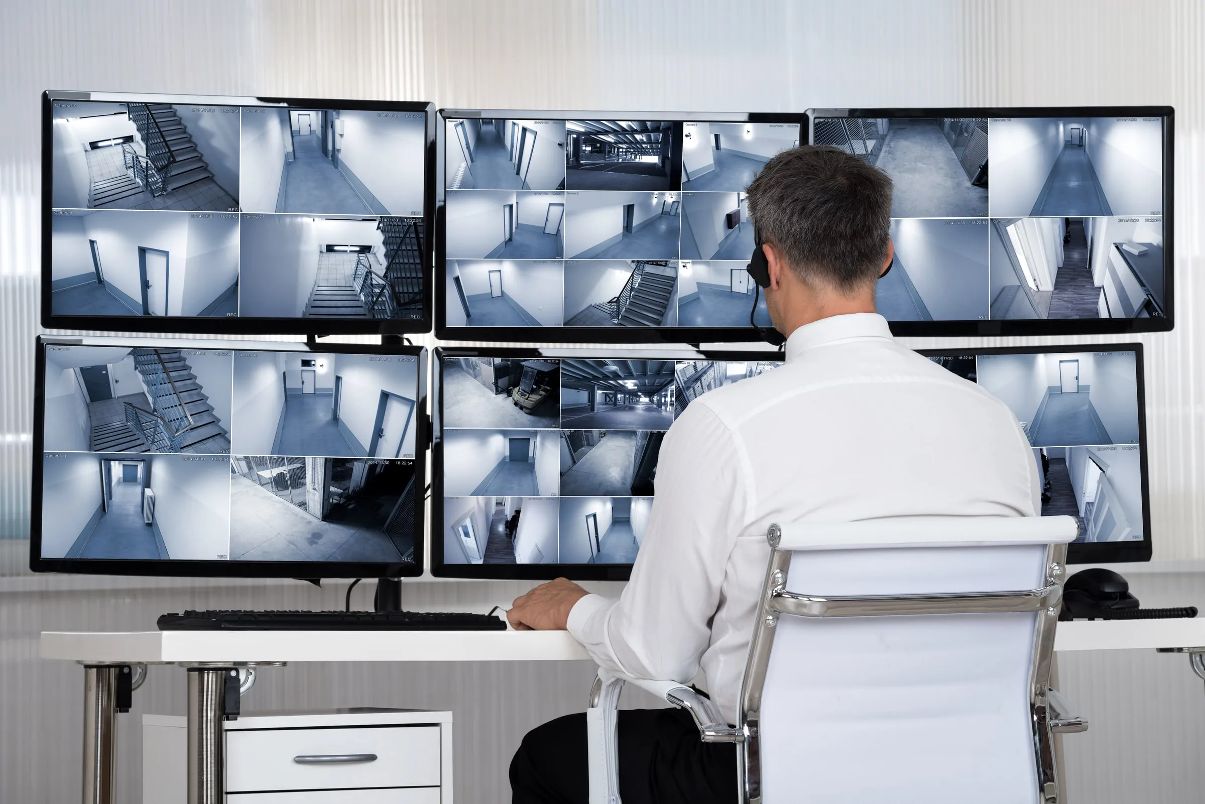 Man in a white shirt seen from behind at a bank of monitors showing stairwells, hallways and a parking garage on split-screen camera feeds