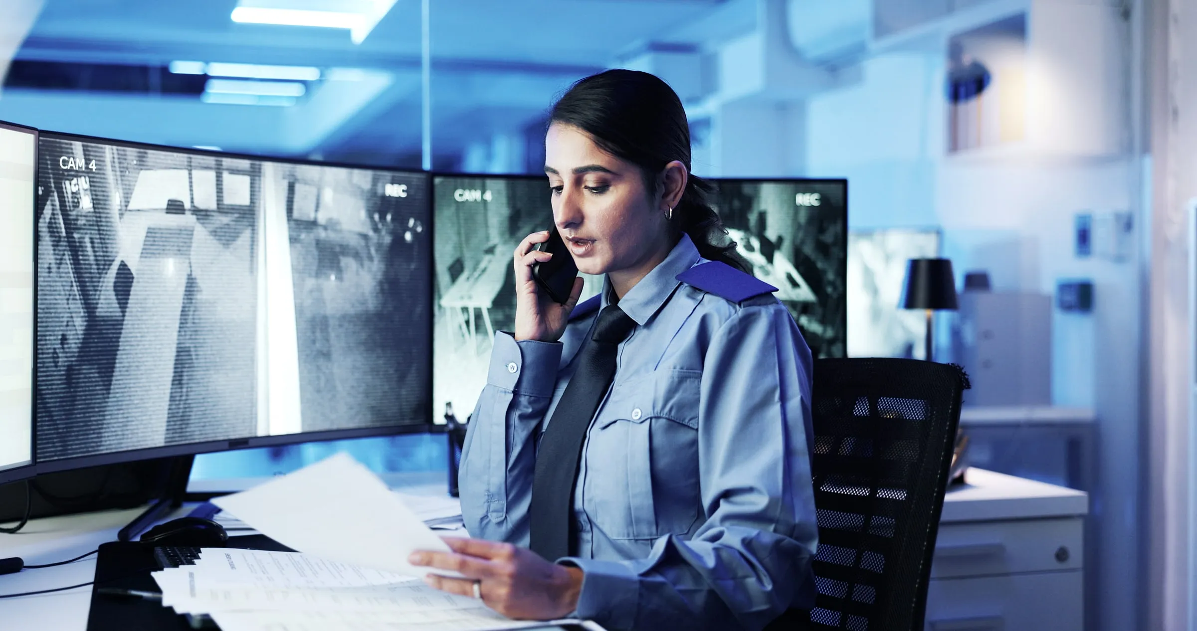 Uniformed officer at a control desk with camera monitors speaks on the phone while reviewing paperwork