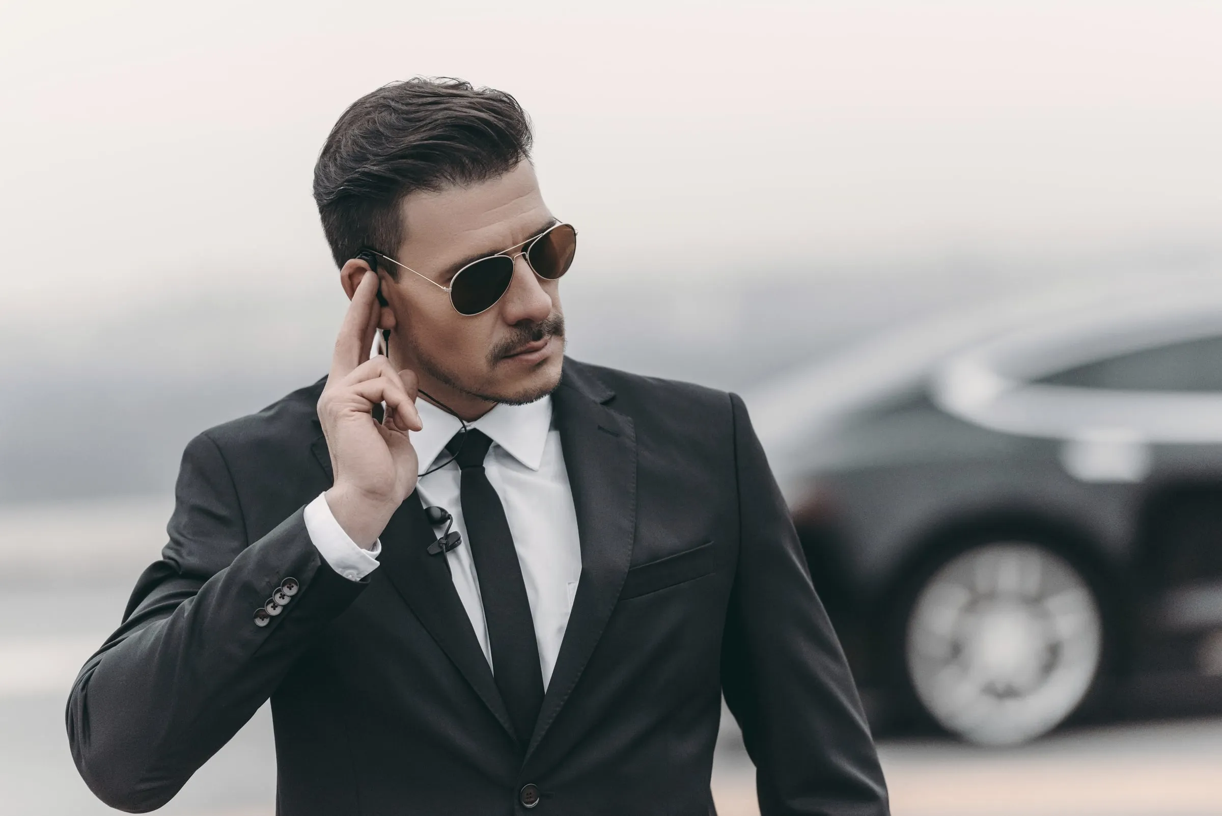 Man in a dark suit and sunglasses adjusts an earpiece while standing beside a black car