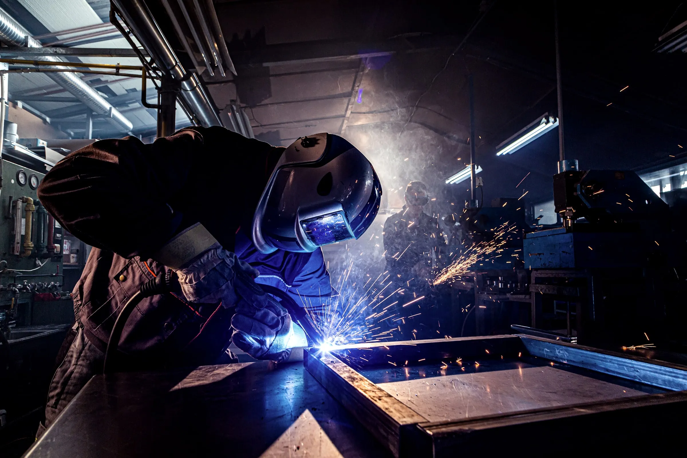 Welder in a face shield and gloves cutting metal in a dark workshop, sparks flying as a second masked worker looks on in the background