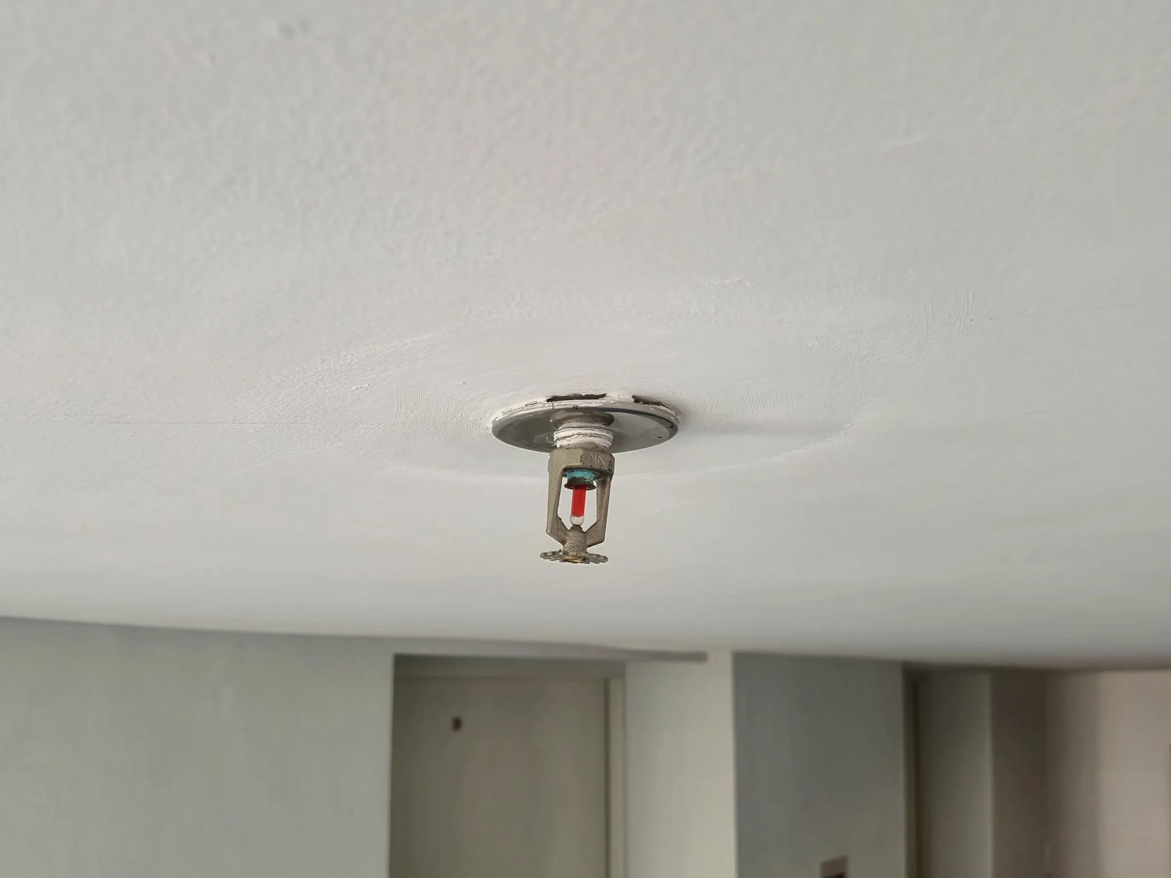 Chrome pendant sprinkler head with a red glass bulb hanging from a white ceiling above a corridor with two doors