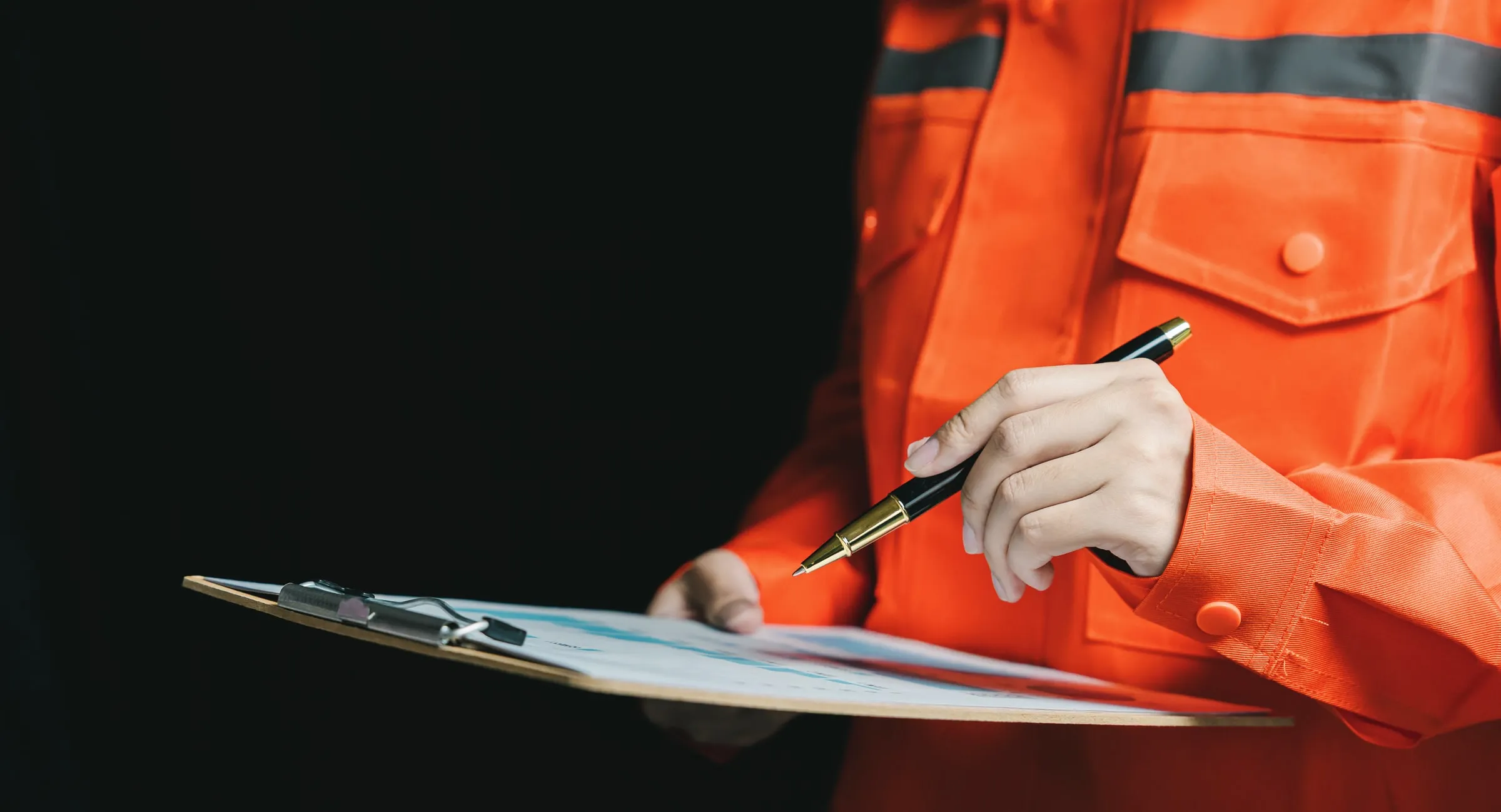 Person in an orange high-visibility jacket holding a clipboard with a pen, pointing at a printed report