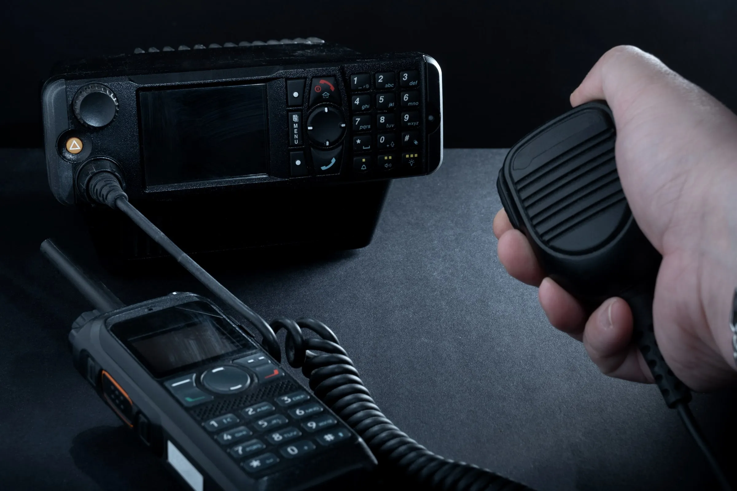 Two-way radio base station beside a handheld unit and a shoulder microphone, on a dark desk in low light