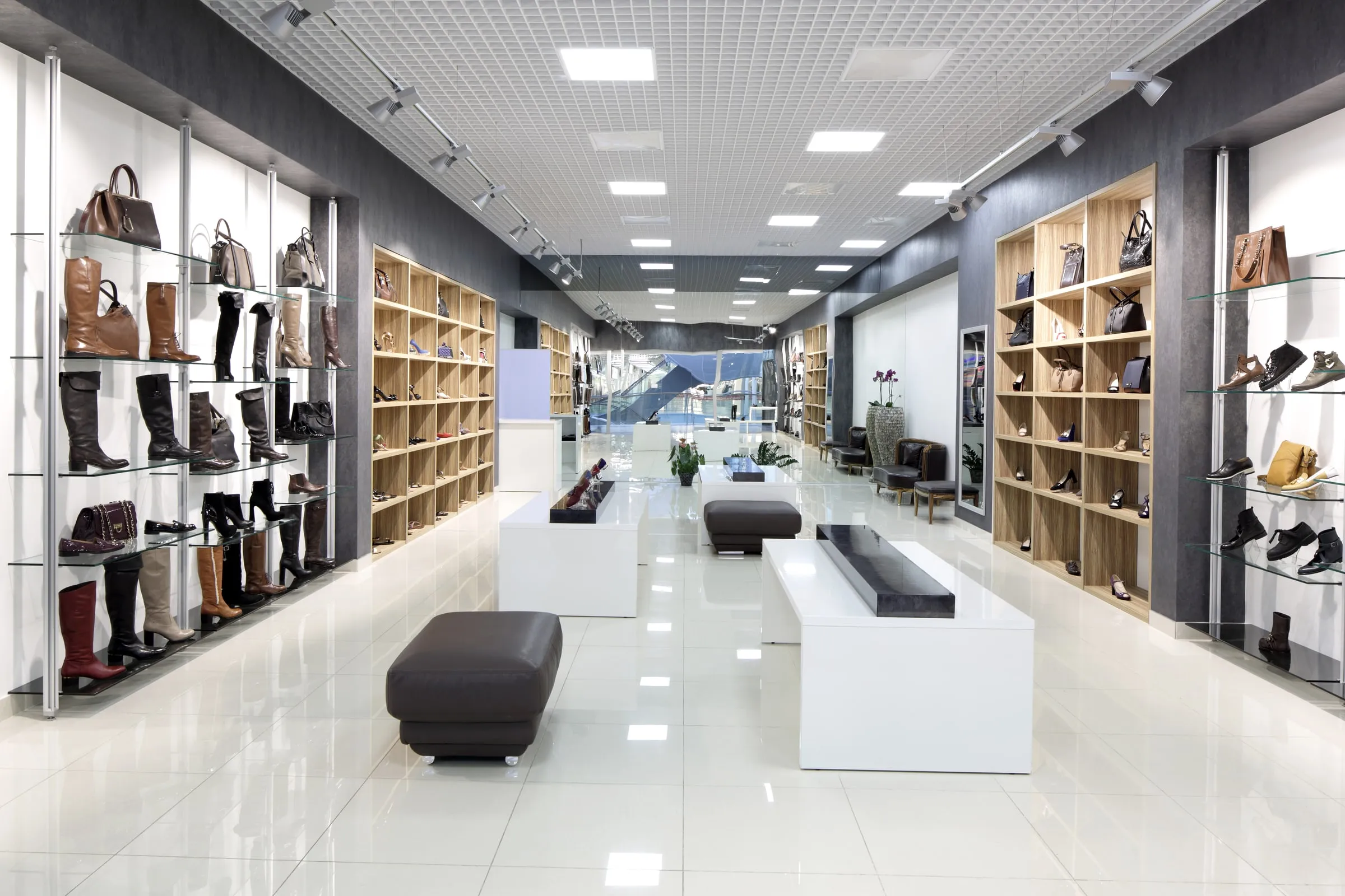 Interior of an upscale boutique retail store with glass shelving of shoes and handbags lining both walls under track lighting