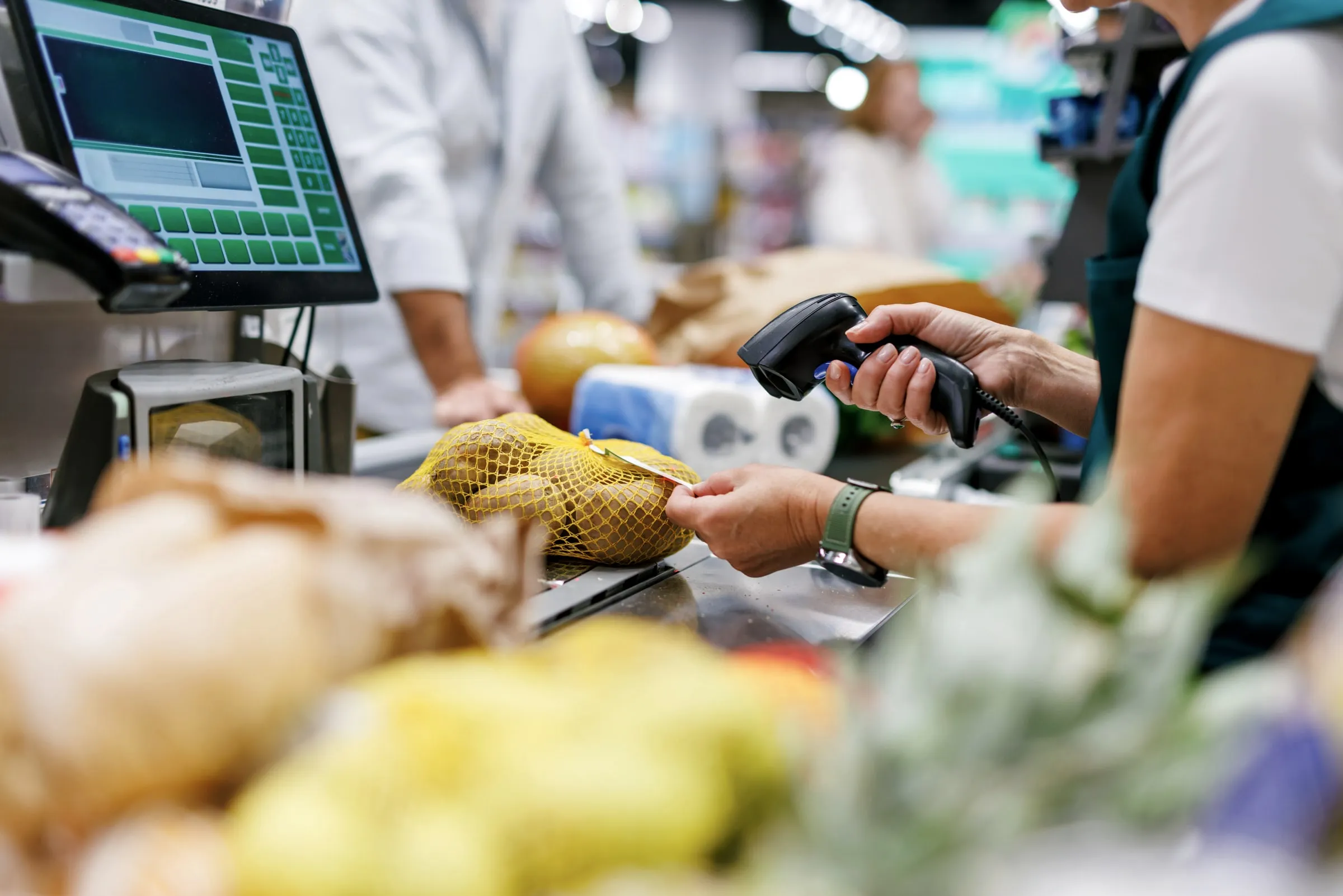 Grocery store checkout close-up of a cashier's hands scanning a mesh bag of potatoes beside a point-of-sale screen
