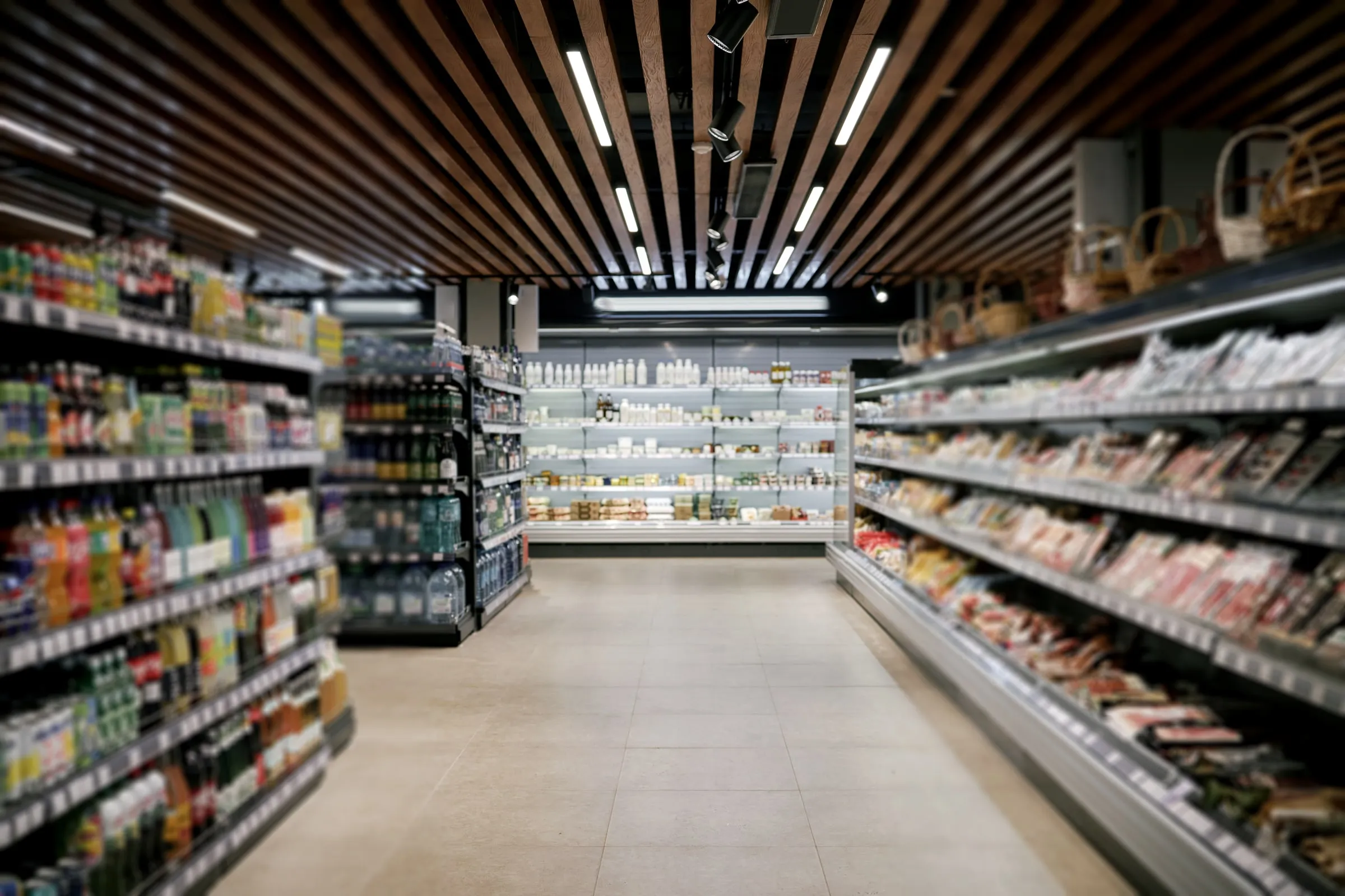 Empty grocery store aisle with beverage shelves on one side and a refrigerated deli case on the other under wood-slat ceiling lighting