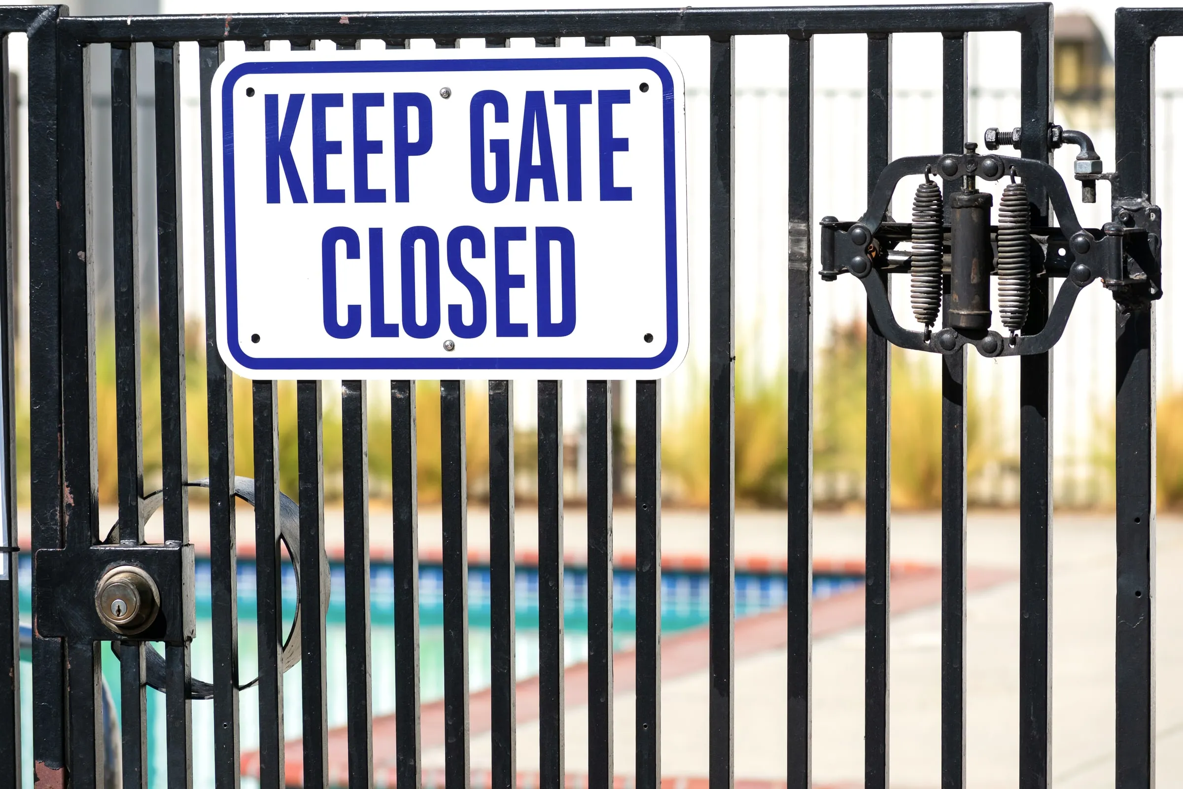 Keep Gate Closed sign mounted on a black iron pool fence, with a swimming pool visible through the bars