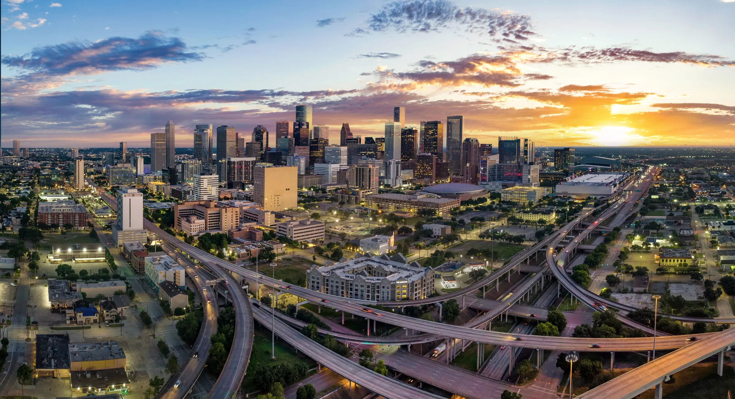 Aerial view of the Grand Parkway and Katy Freeway stack interchange under a partly cloudy sky, with flat green land and retail rooftops in the distance