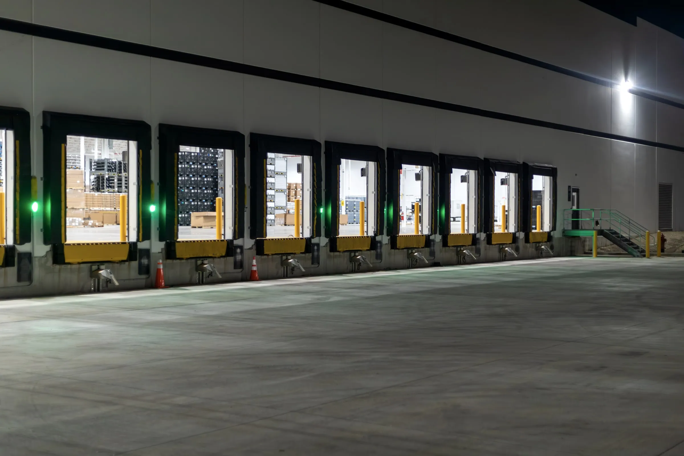 Row of illuminated loading dock bays at a warehouse at night, doors open with pallets of goods visible inside, green dock lights lit