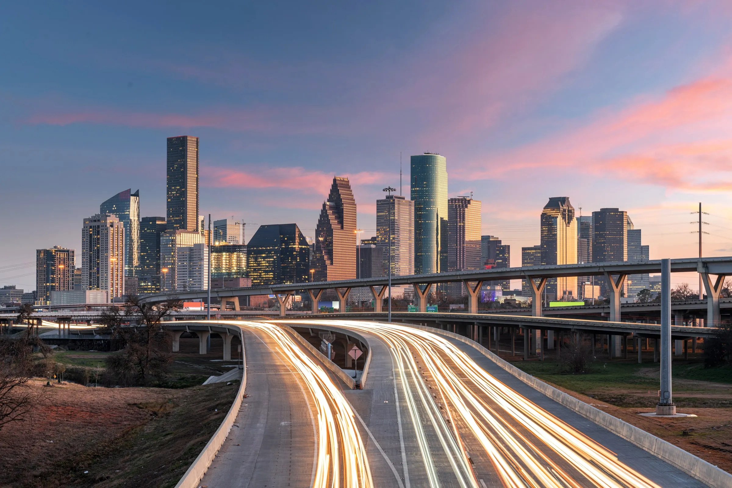 Elevated highway interchange with long-exposure light trails from traffic leading toward the downtown Houston skyline at dusk