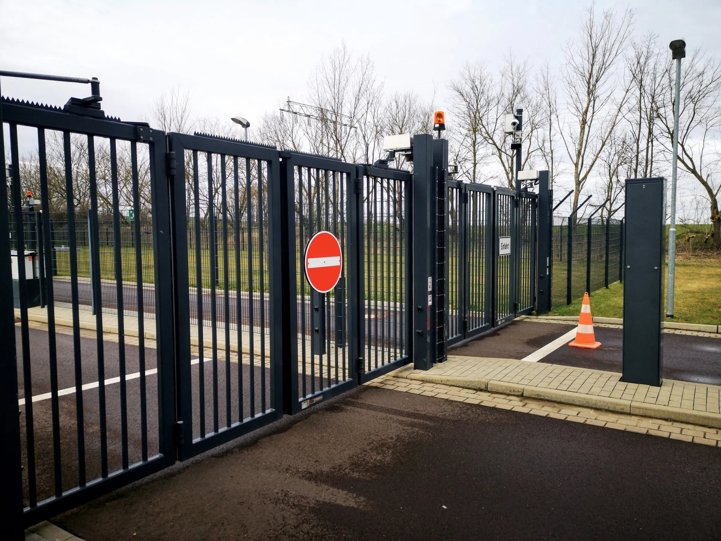 Black metal automatic gate barrier with a red do-not-enter sign, security cameras and an orange traffic cone at a controlled entrance