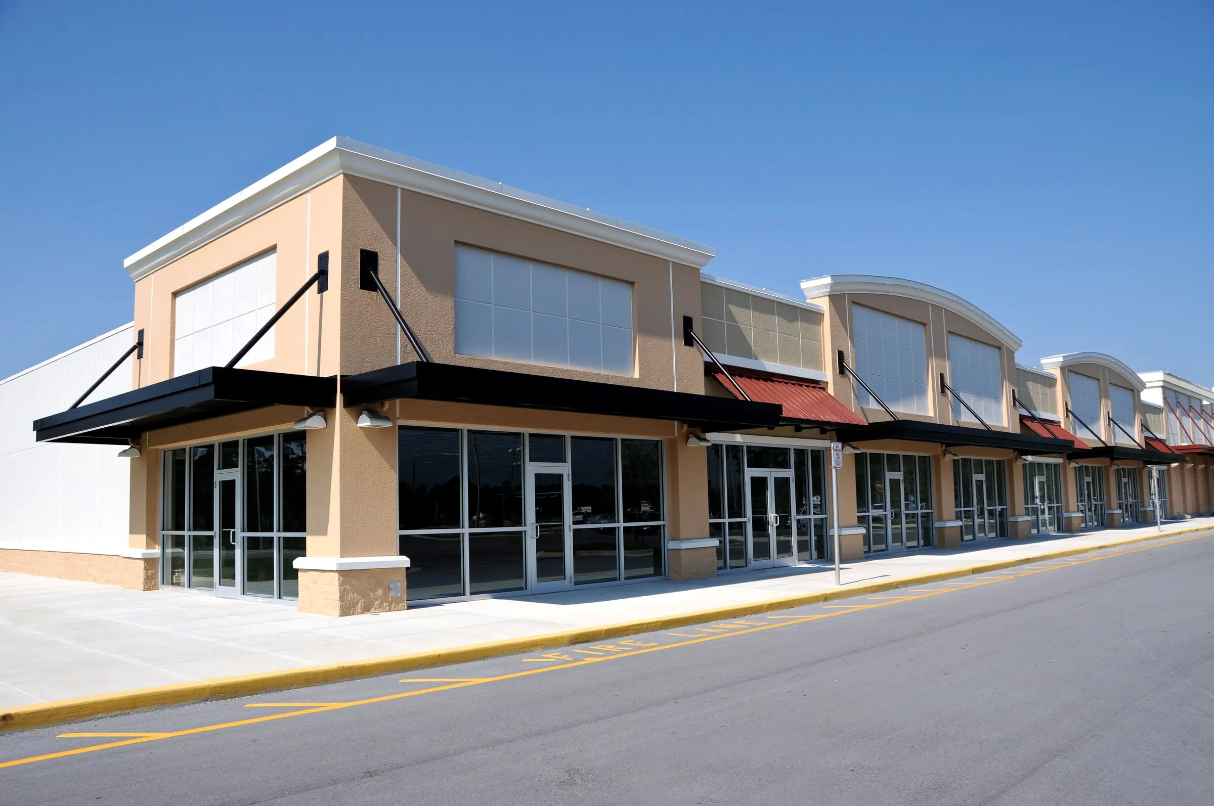 Single-storey retail strip center with tan stucco, black awnings and glass storefronts beside an empty parking lot under a clear sky
