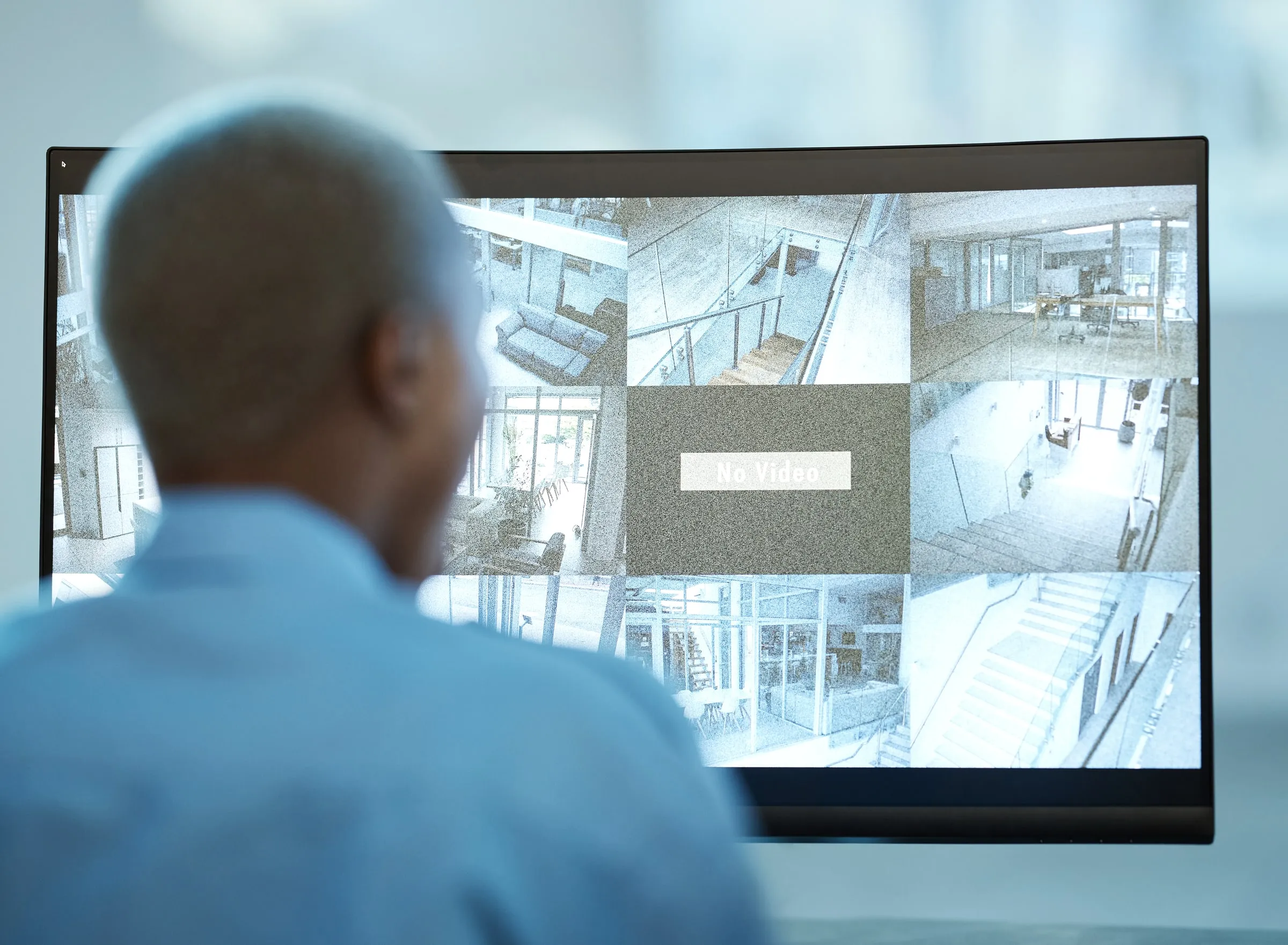 Man viewed from behind watching a wall-mounted monitor split into camera feeds of a building's stairwells, lobby and offices