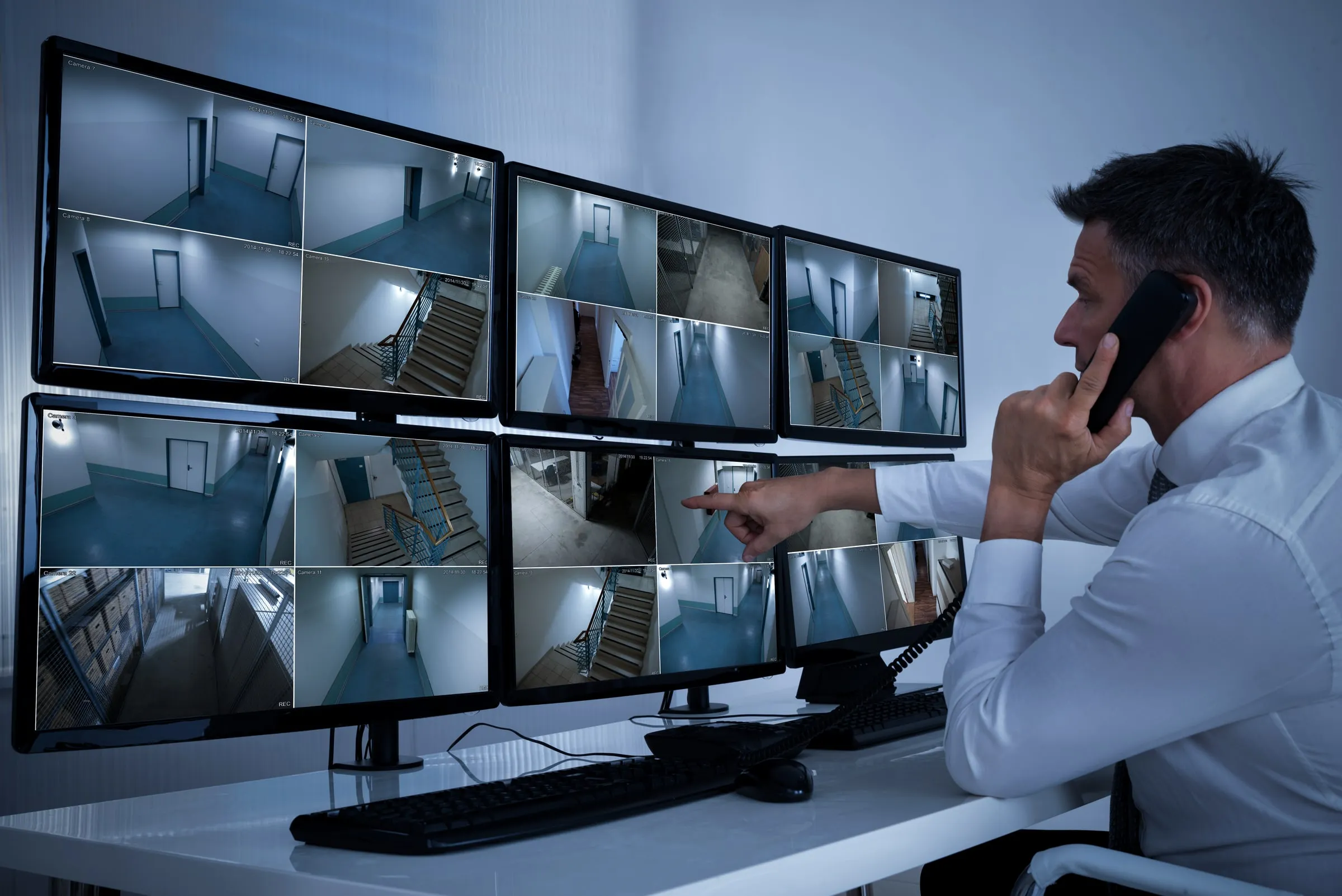 Man in a white shirt on the phone, pointing at a bank of monitors showing stairwells, corridors and doors