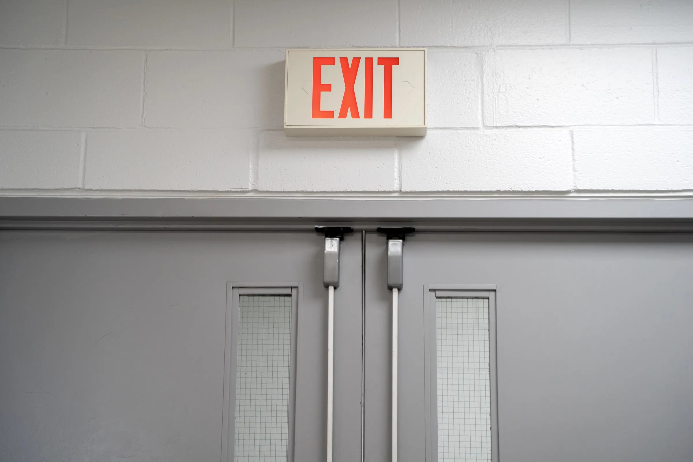 Illuminated red EXIT sign above a closed pair of gray double doors with crash bars, in a painted cinder block corridor
