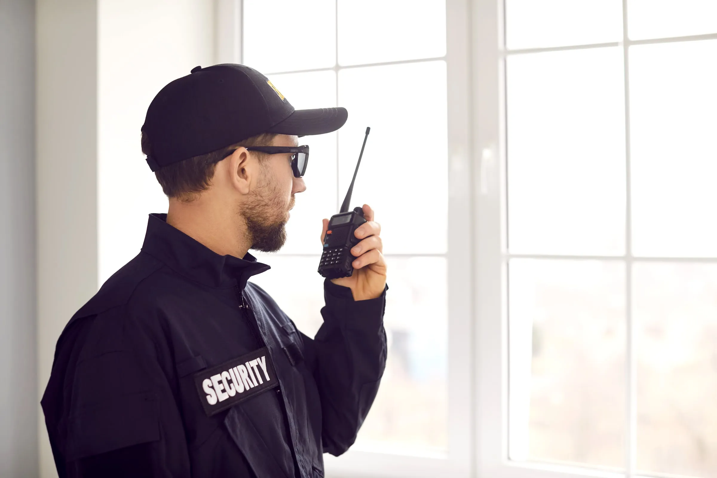 Uniformed security officer holding a two-way radio to speak, standing beside a bright window