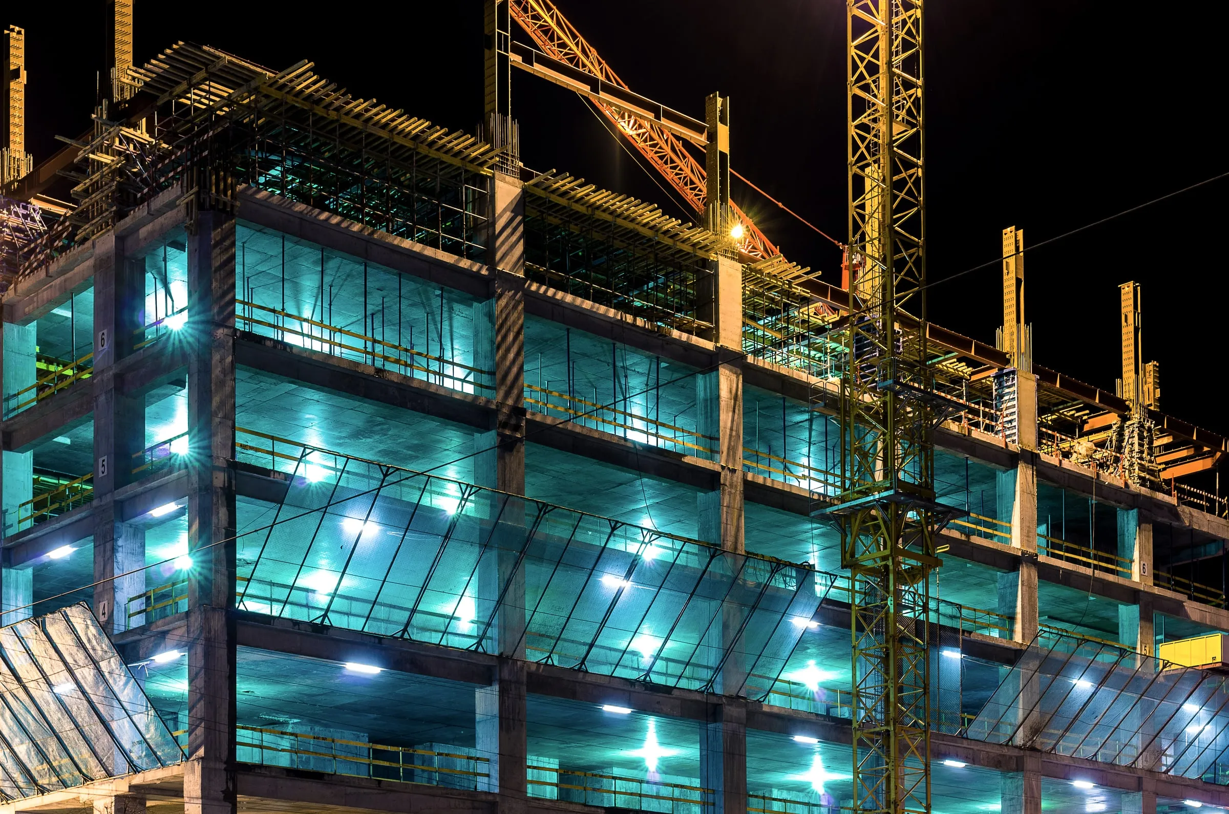 High-rise building under construction at night, lit from within, with a crane rising against a dark sky