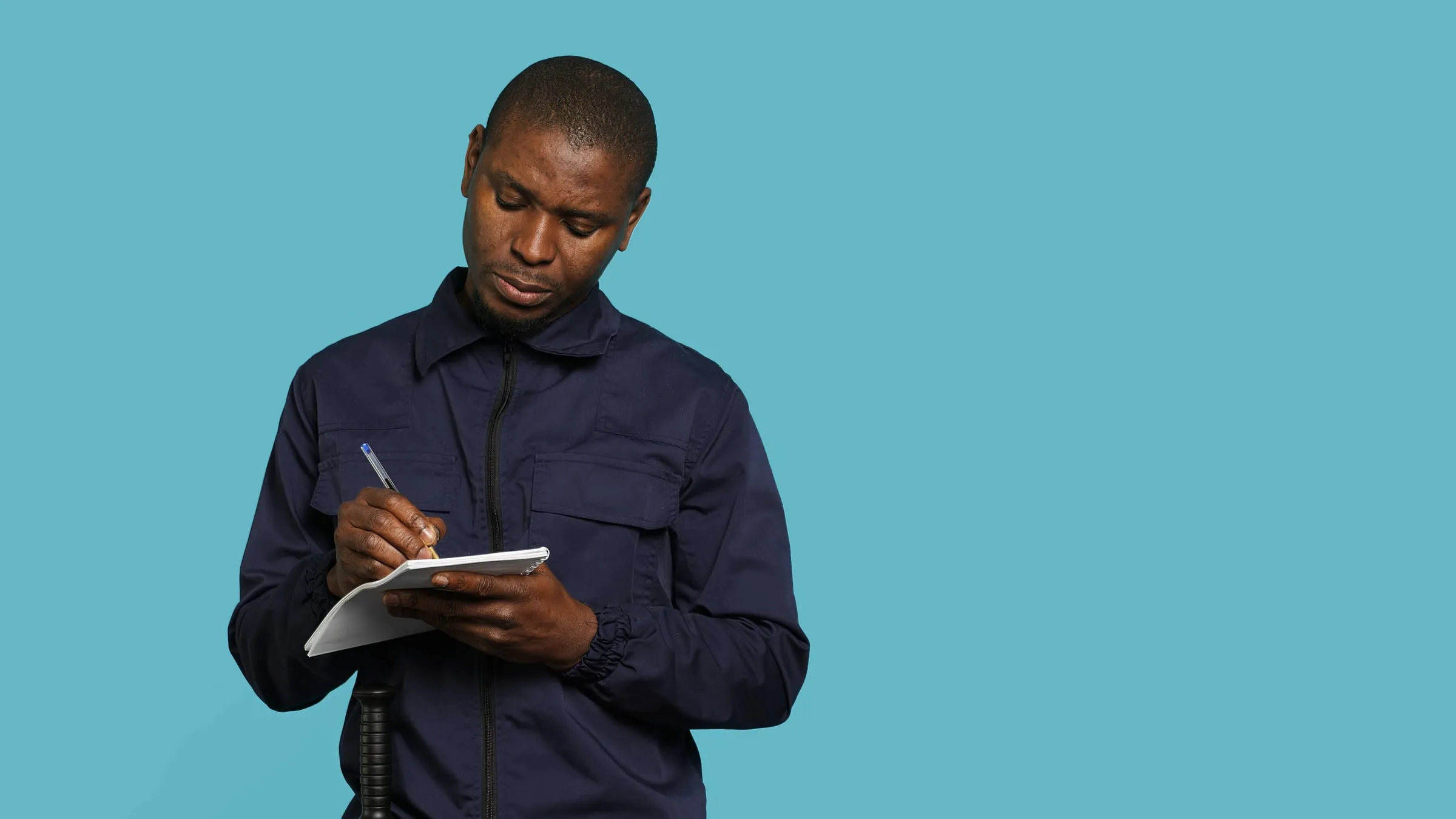 Man in navy coveralls writing on a small spiral notepad against a plain blue backdrop