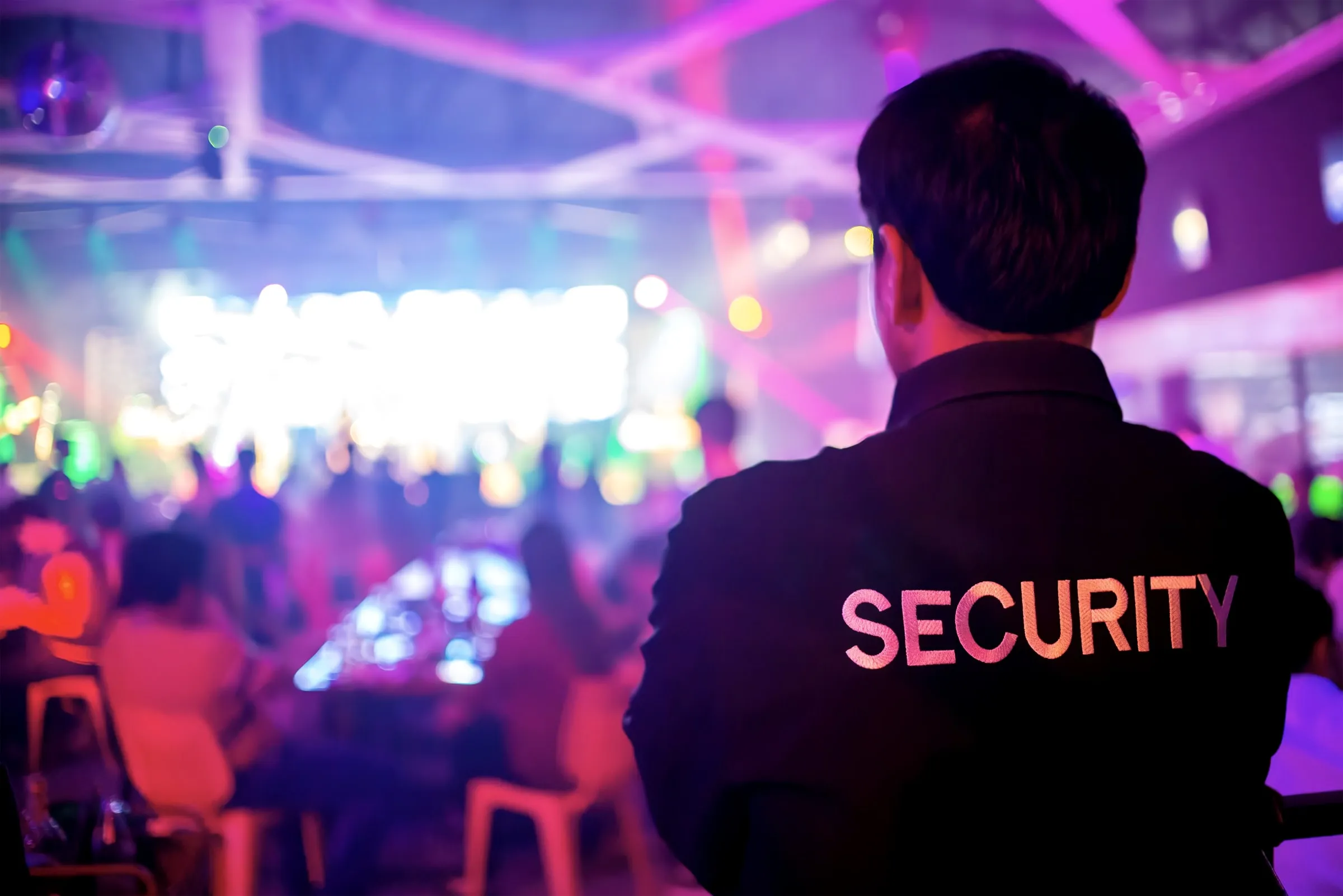 Rear view of a uniformed security officer wearing a jacket marked SECURITY, standing watch over a crowded indoor event under colored lights