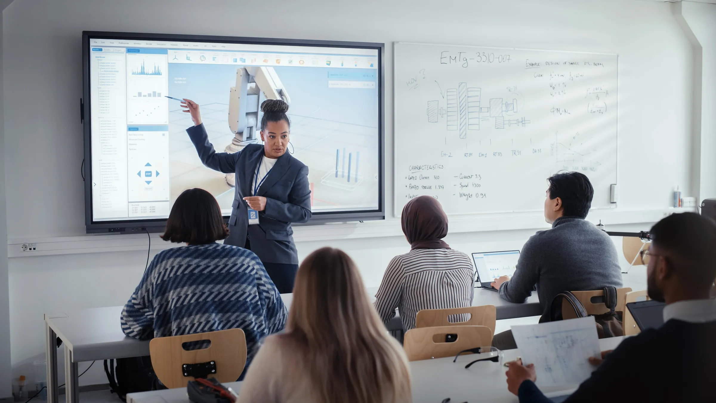 Instructor pointing to a diagram on a large screen while seated trainees take notes, with a technical whiteboard behind
