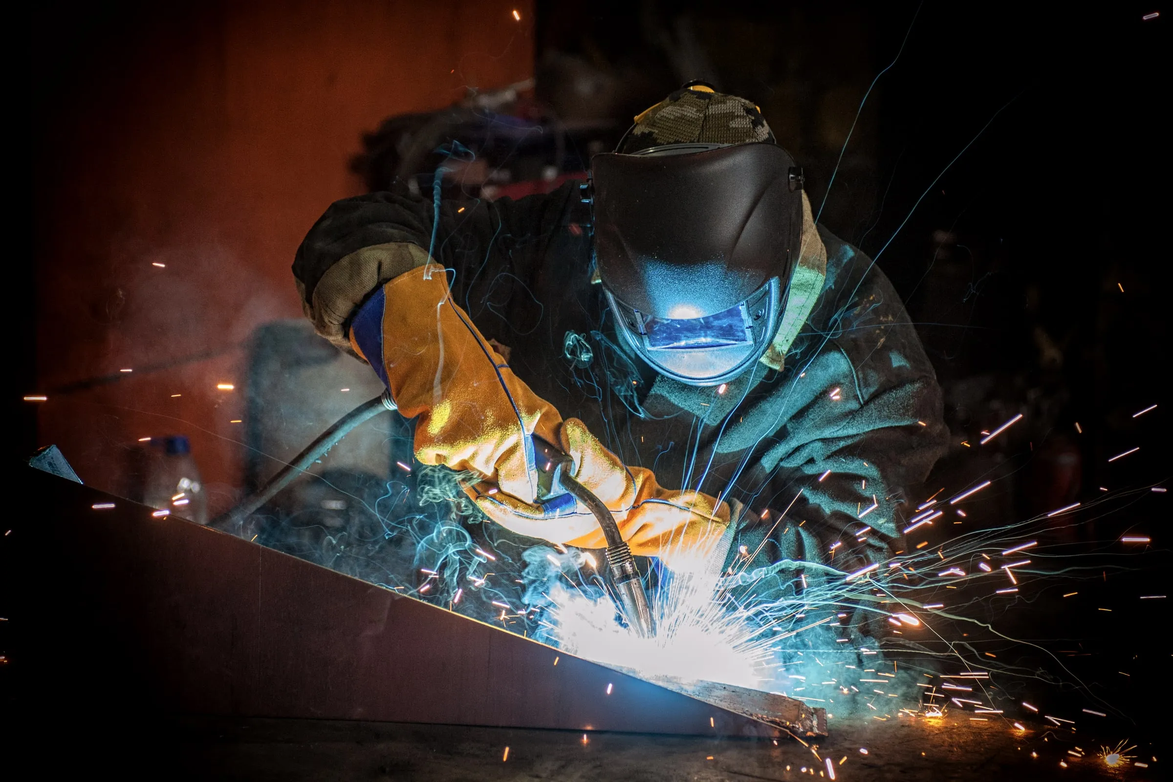 Welder in a face shield and heavy yellow gloves cutting an angled steel beam at close range, sparks arcing across a dark workshop