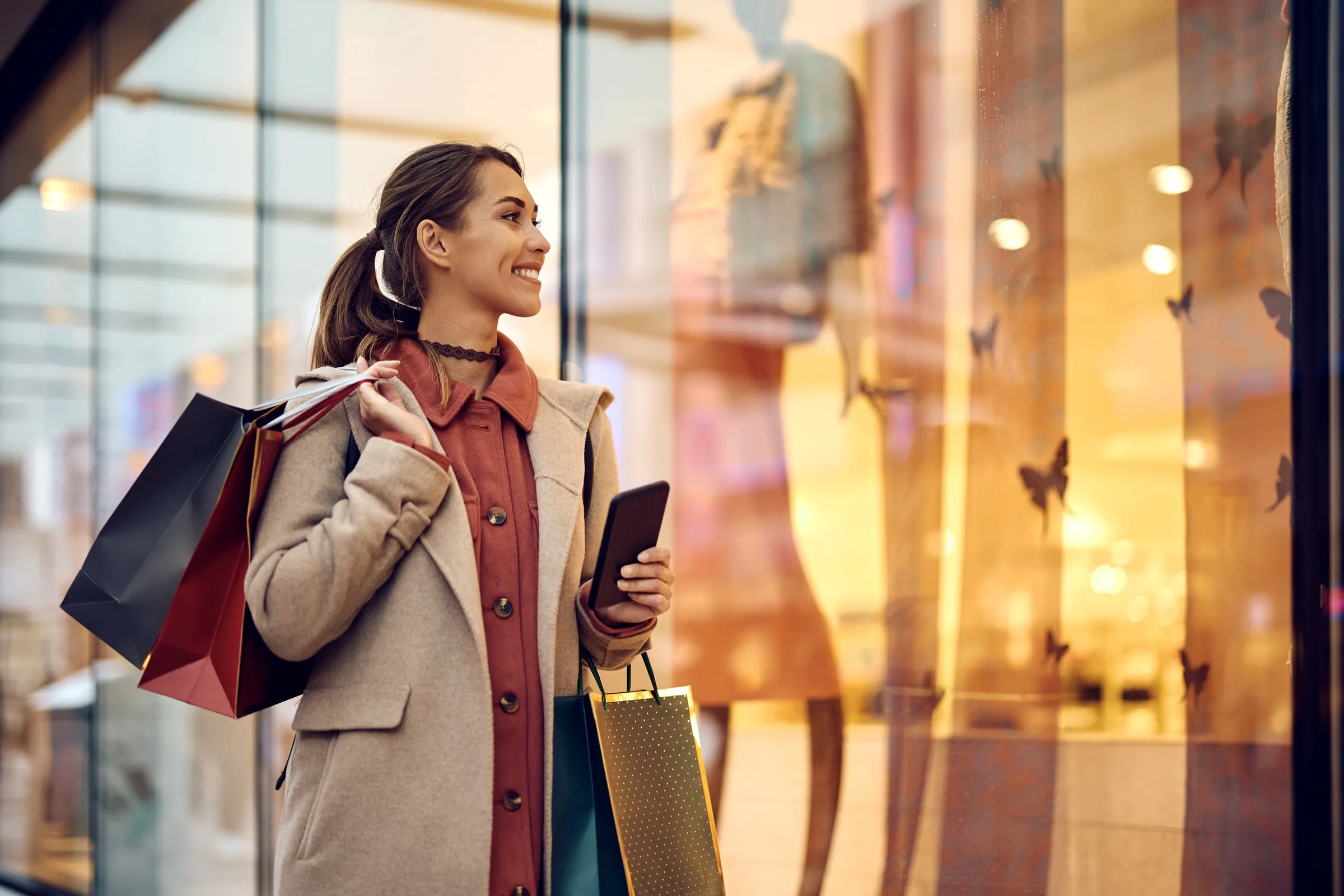 Woman carrying shopping bags smiles while looking at a lit storefront window display in a shopping center