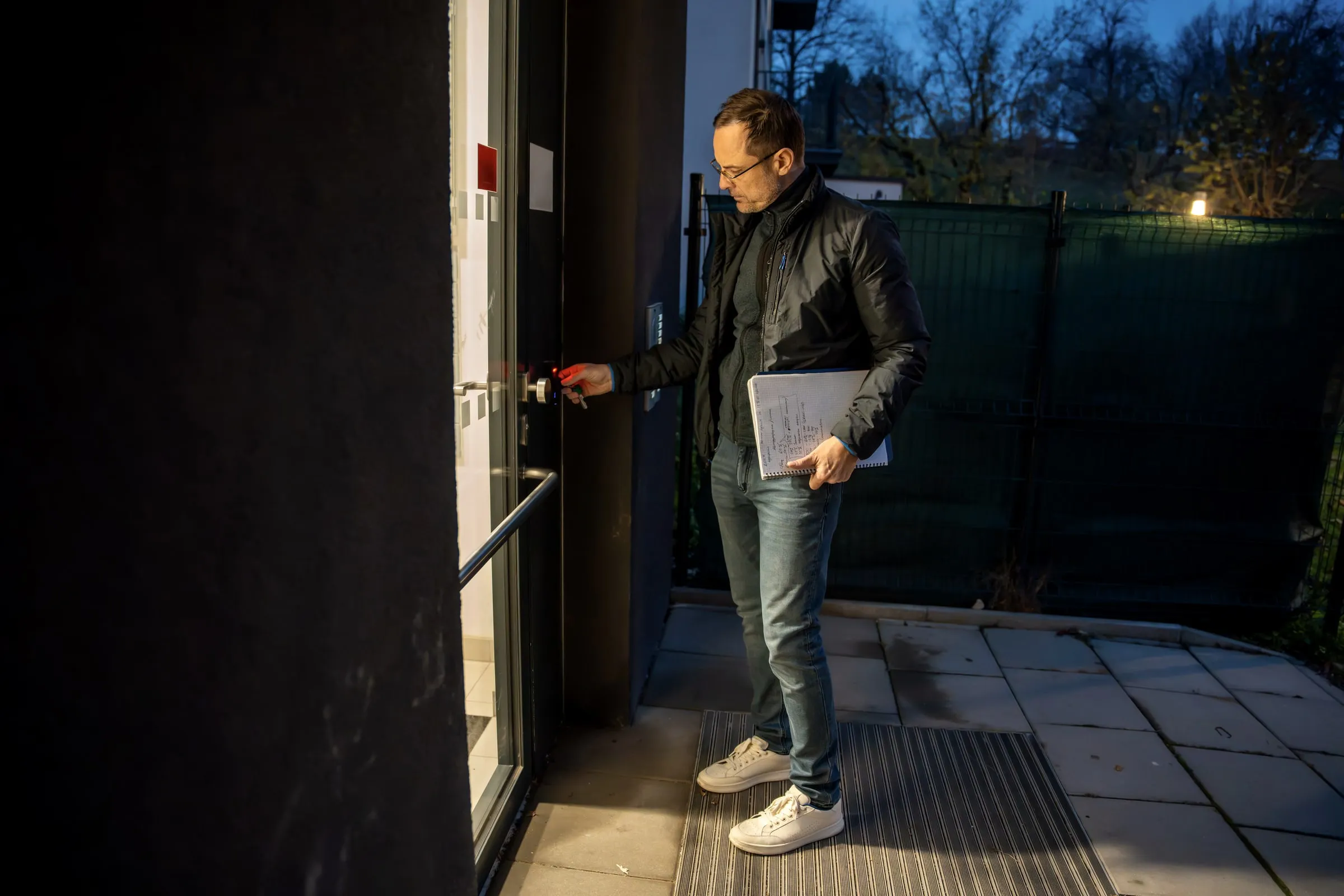 Man in a dark jacket holds a notebook while checking an electronic door lock at a building entrance at dusk