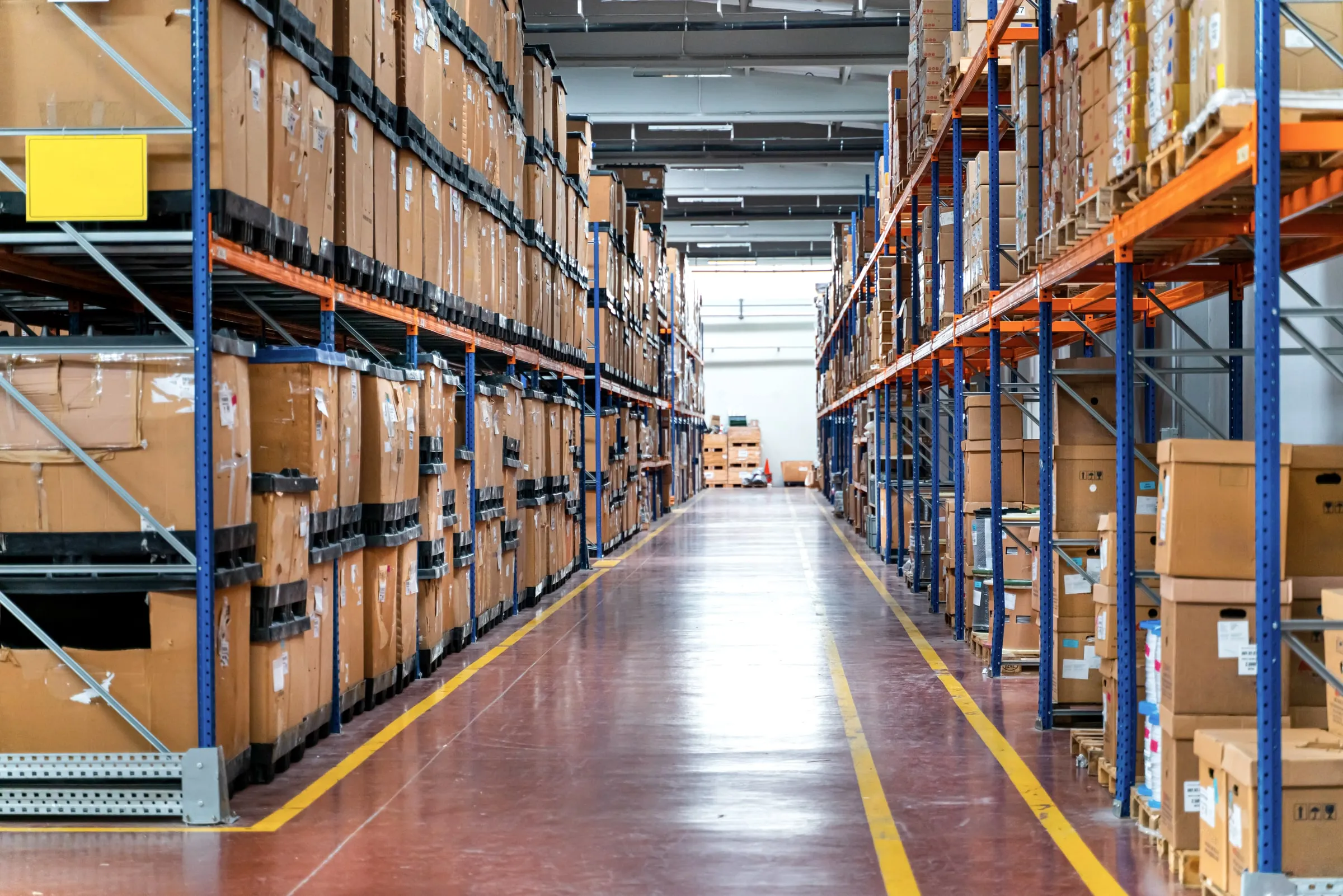 Warehouse aisle between tall pallet racks loaded with boxes, with yellow lines marking the floor