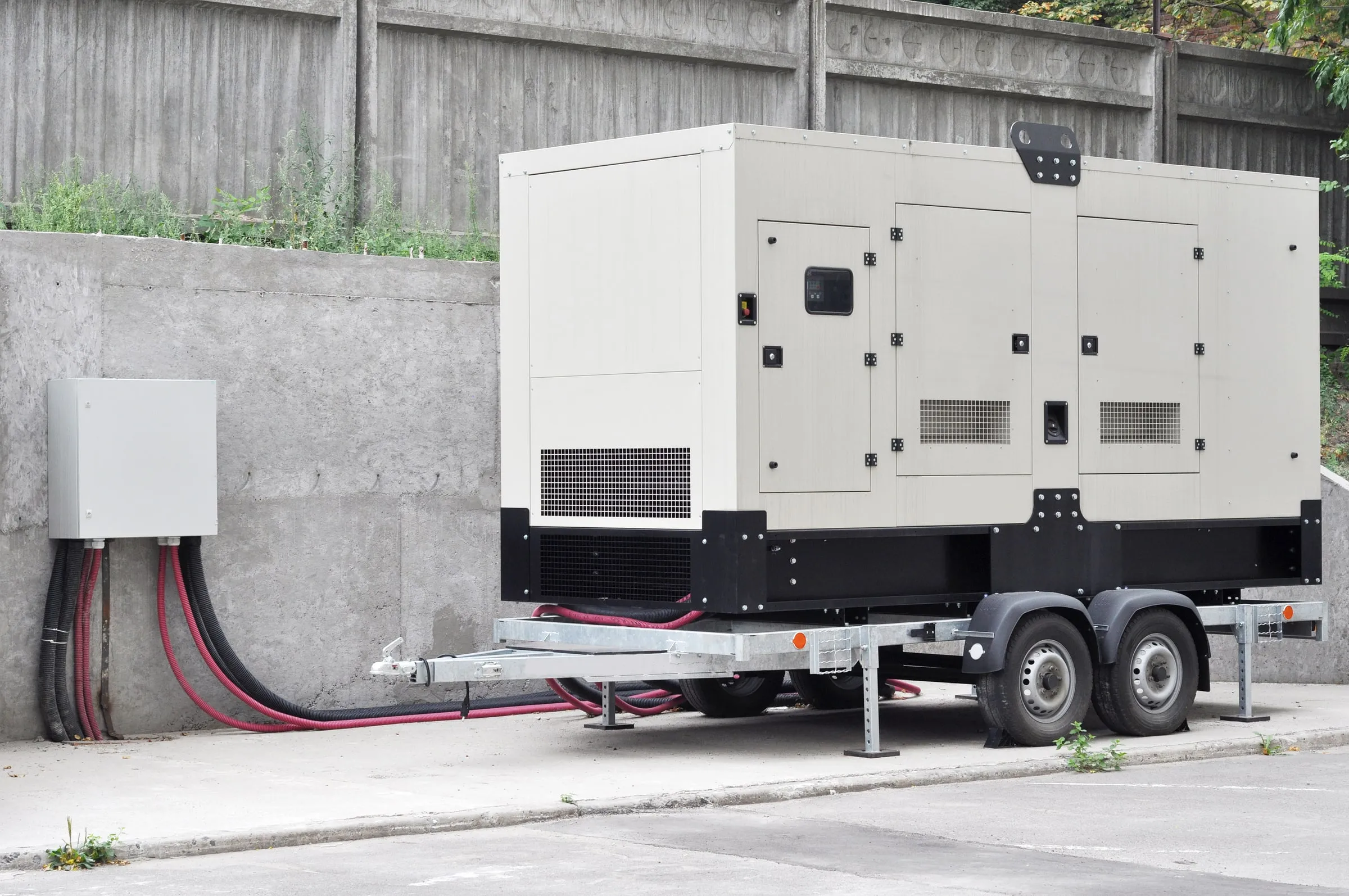 Large towable diesel generator connected by heavy cables to a building's electrical panel against a concrete retaining wall