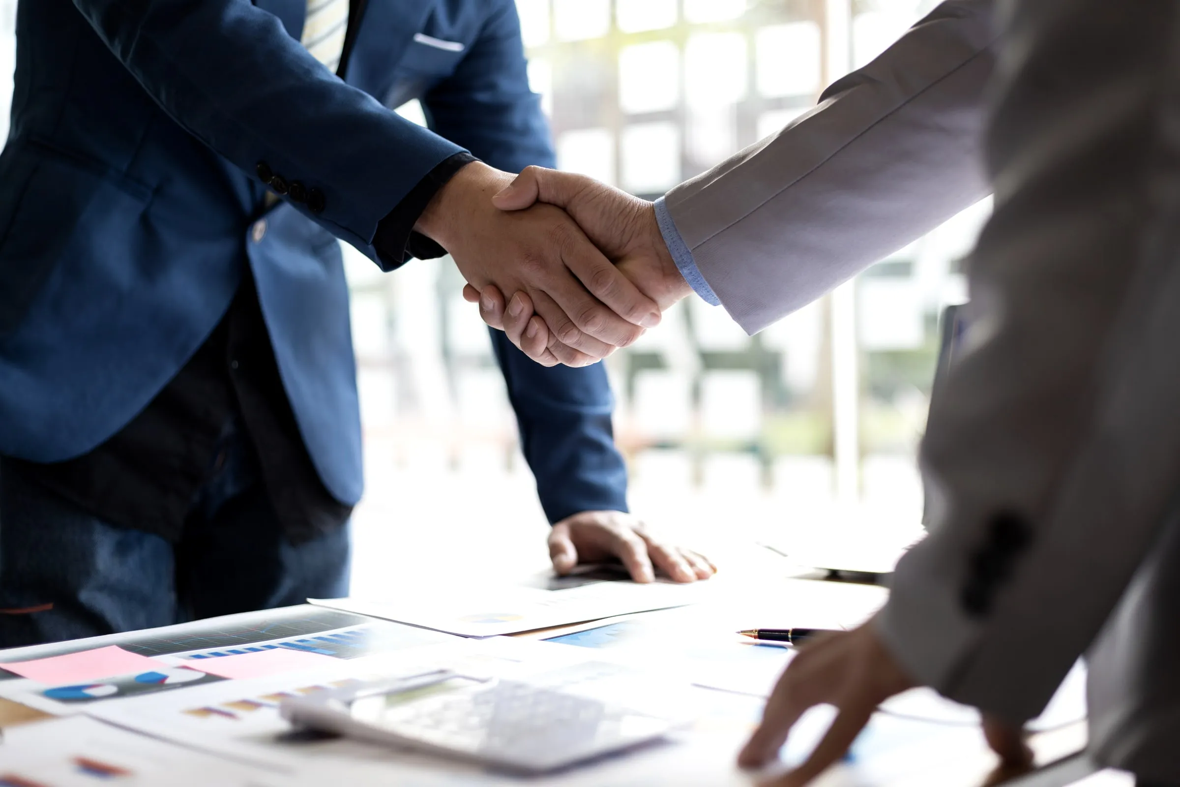 Close-up of two business people in suits shaking hands over a desk covered in charts and a pen