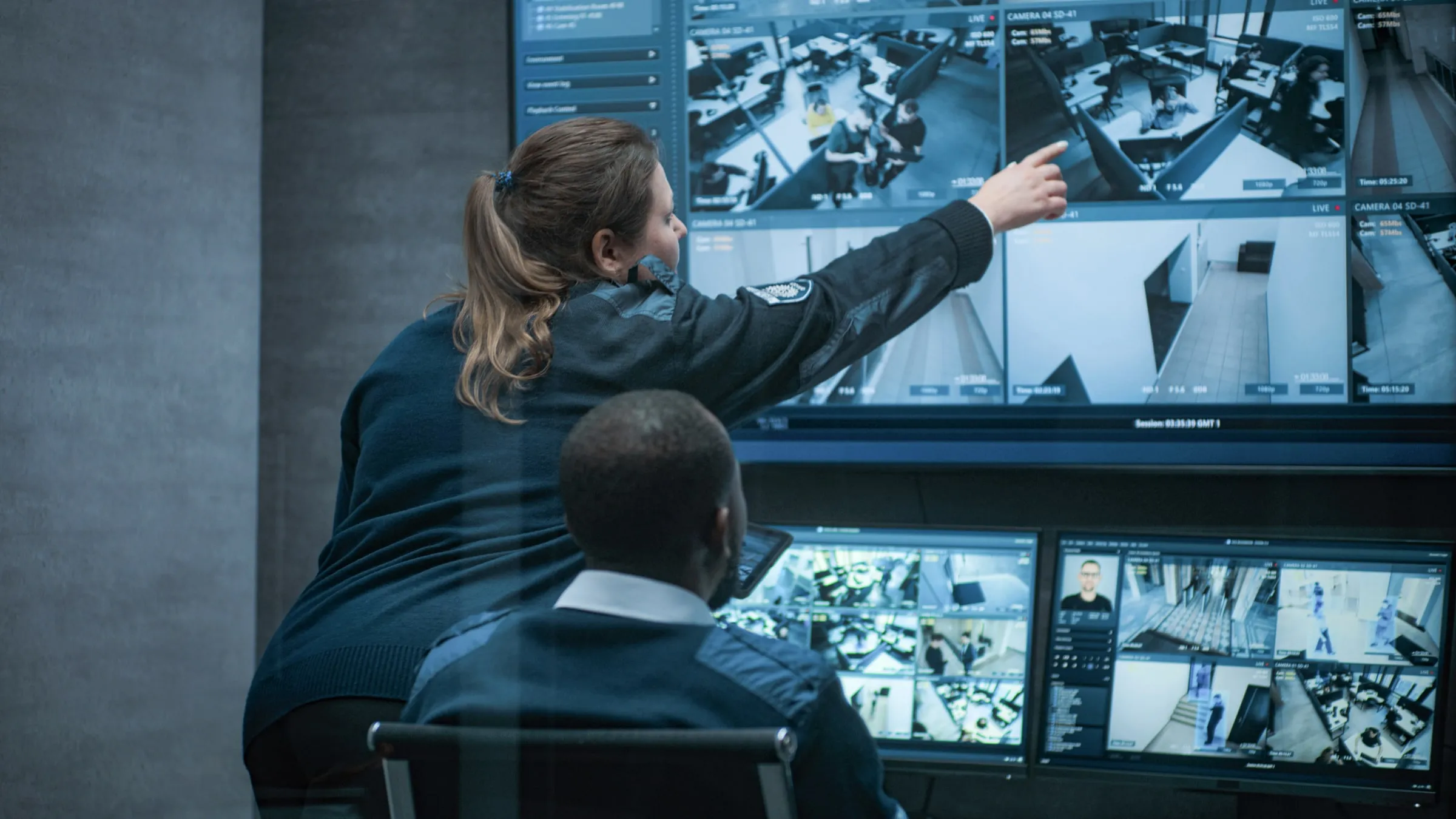 Two uniformed officers in a control room, one pointing at a wall of monitors showing lobbies, corridors and a parking garage