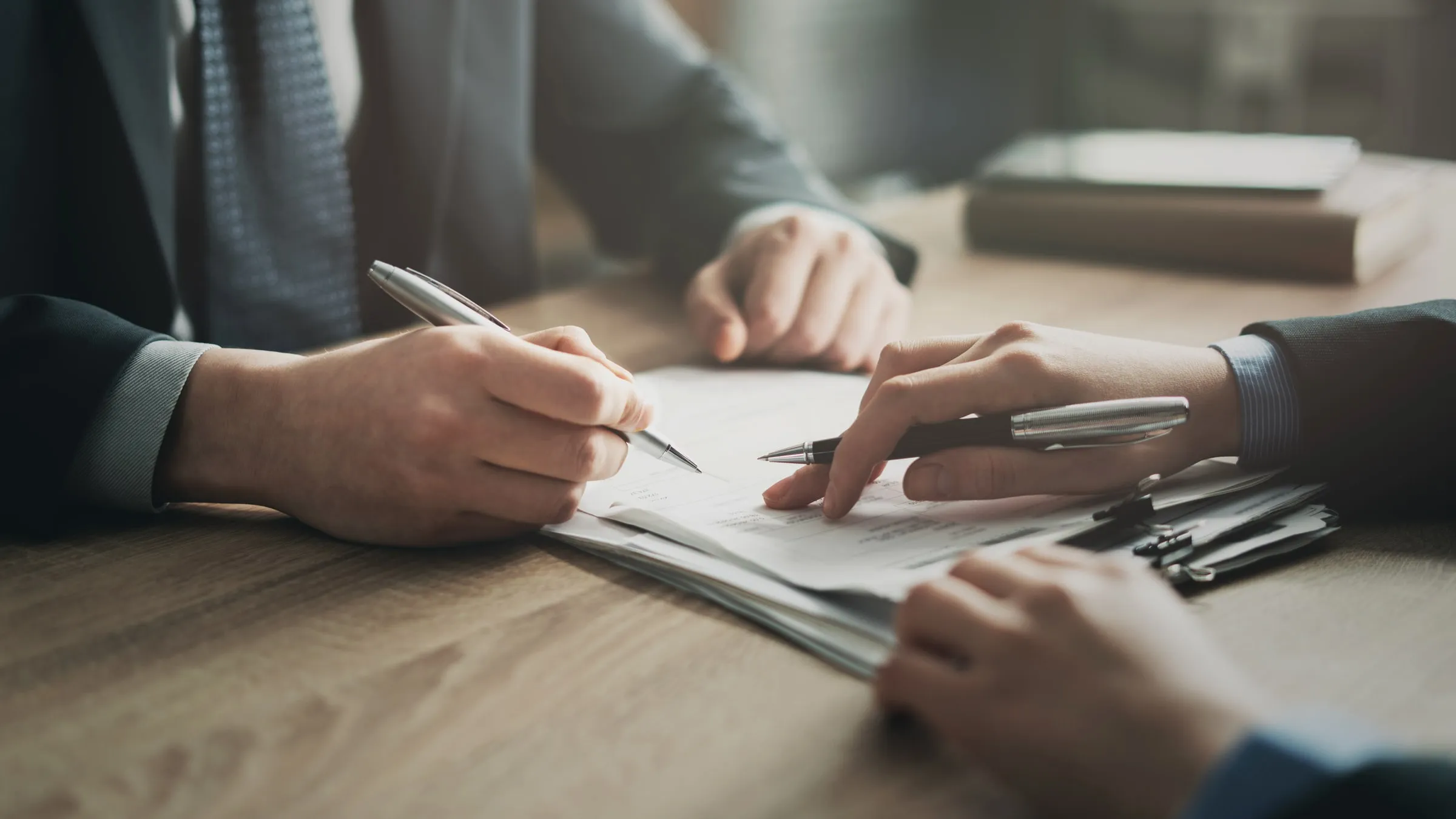 Close-up of two people's hands at a wood table, one signing a document, with keys and a folder alongside the paperwork