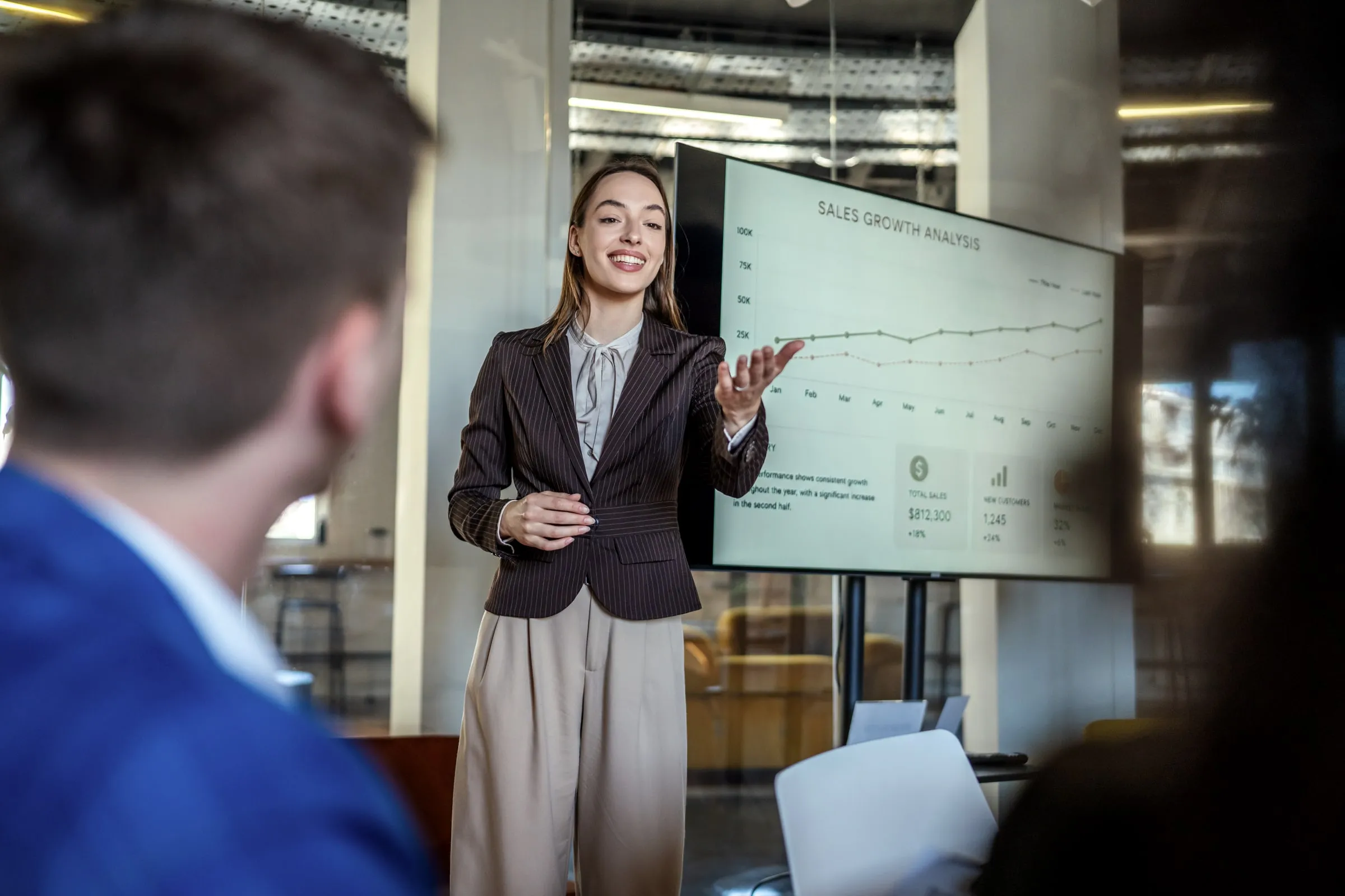 Woman in a pinstripe blazer presenting a sales growth chart on a screen to seated colleagues