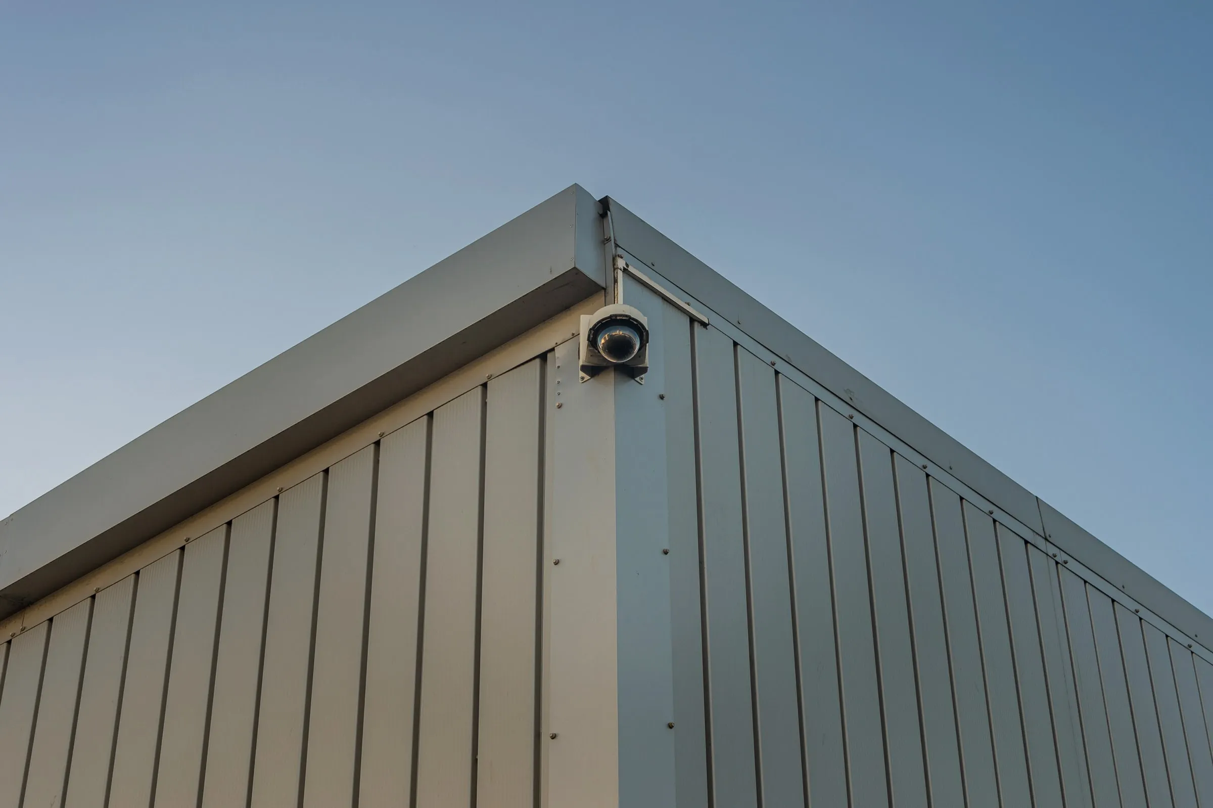 Corner of a metal-sided warehouse building against a clear blue sky, with a dome security camera mounted under the roofline