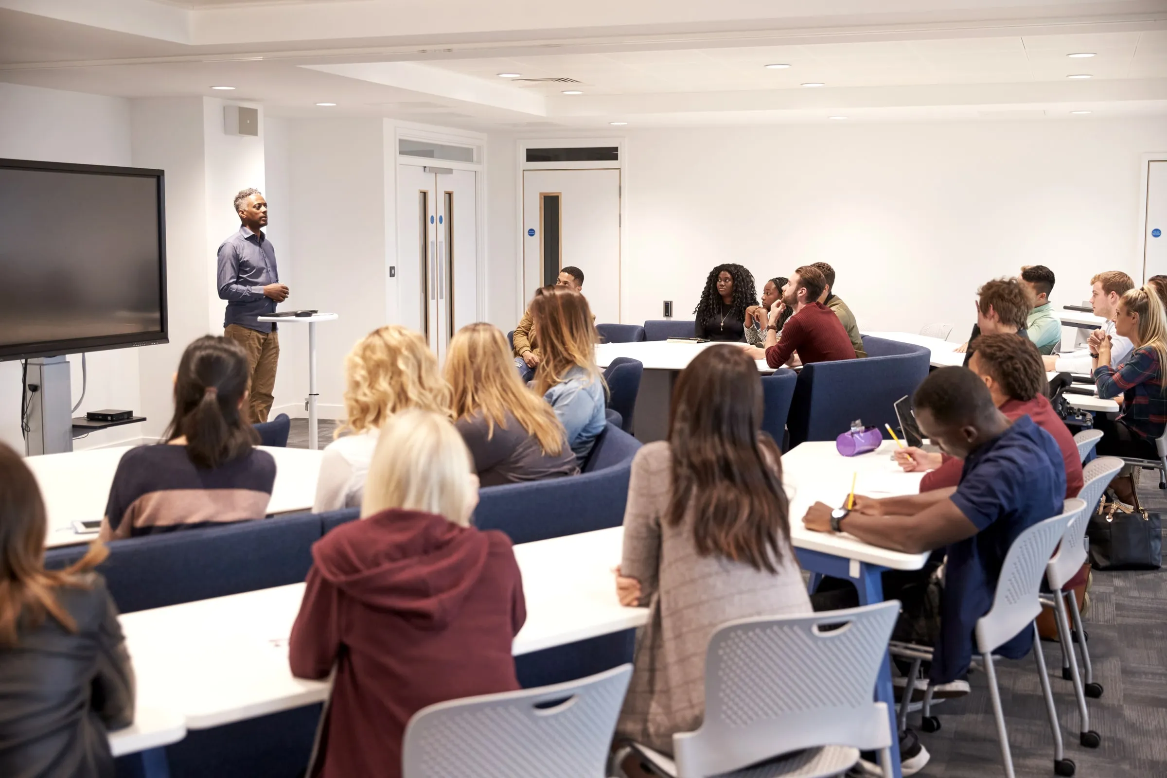 A trainee raises a hand in a lecture hall as an instructor speaks beside a wall-mounted screen and whiteboard