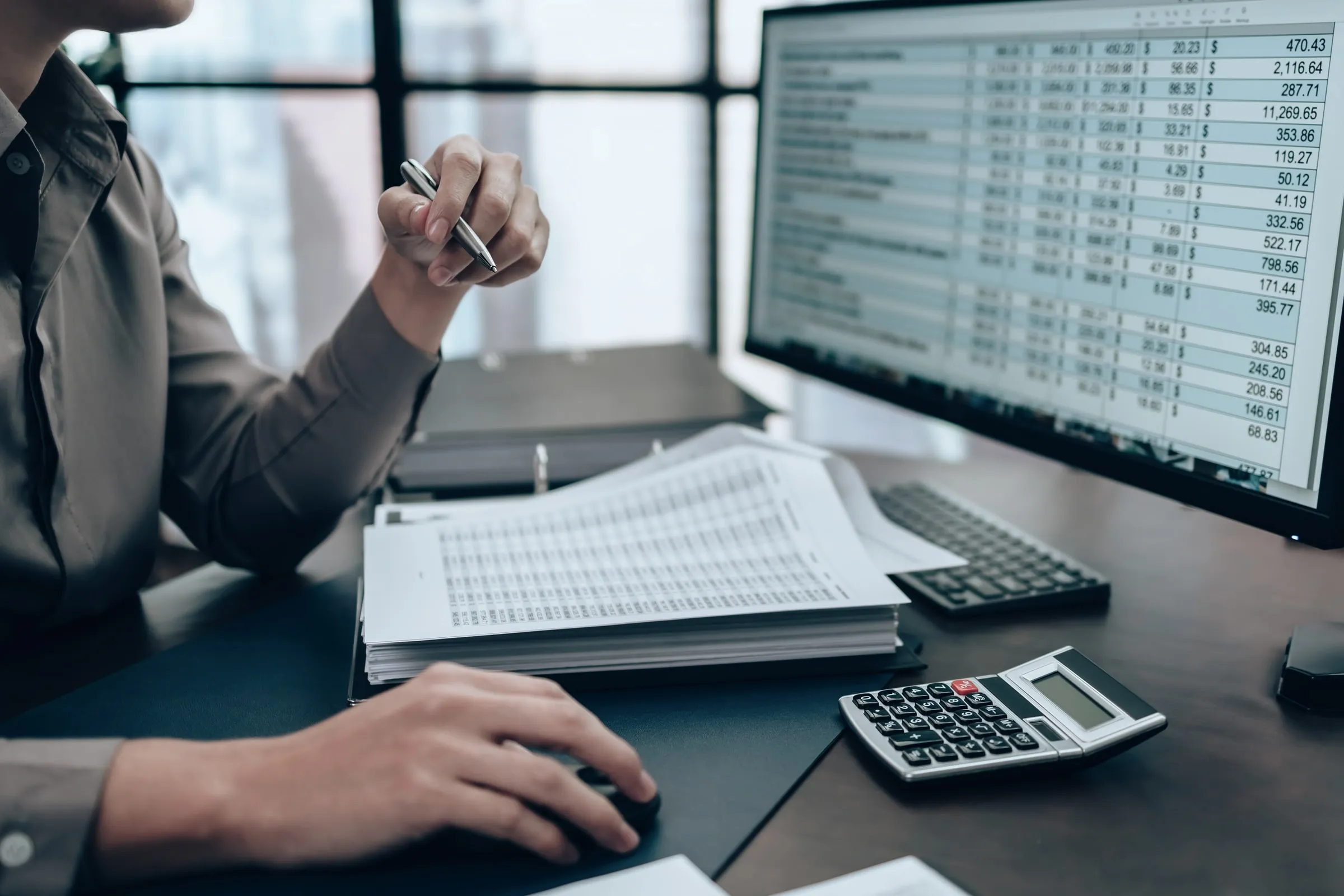 Person at a desk pointing a pen at a spreadsheet on a monitor beside a stack of papers and a calculator