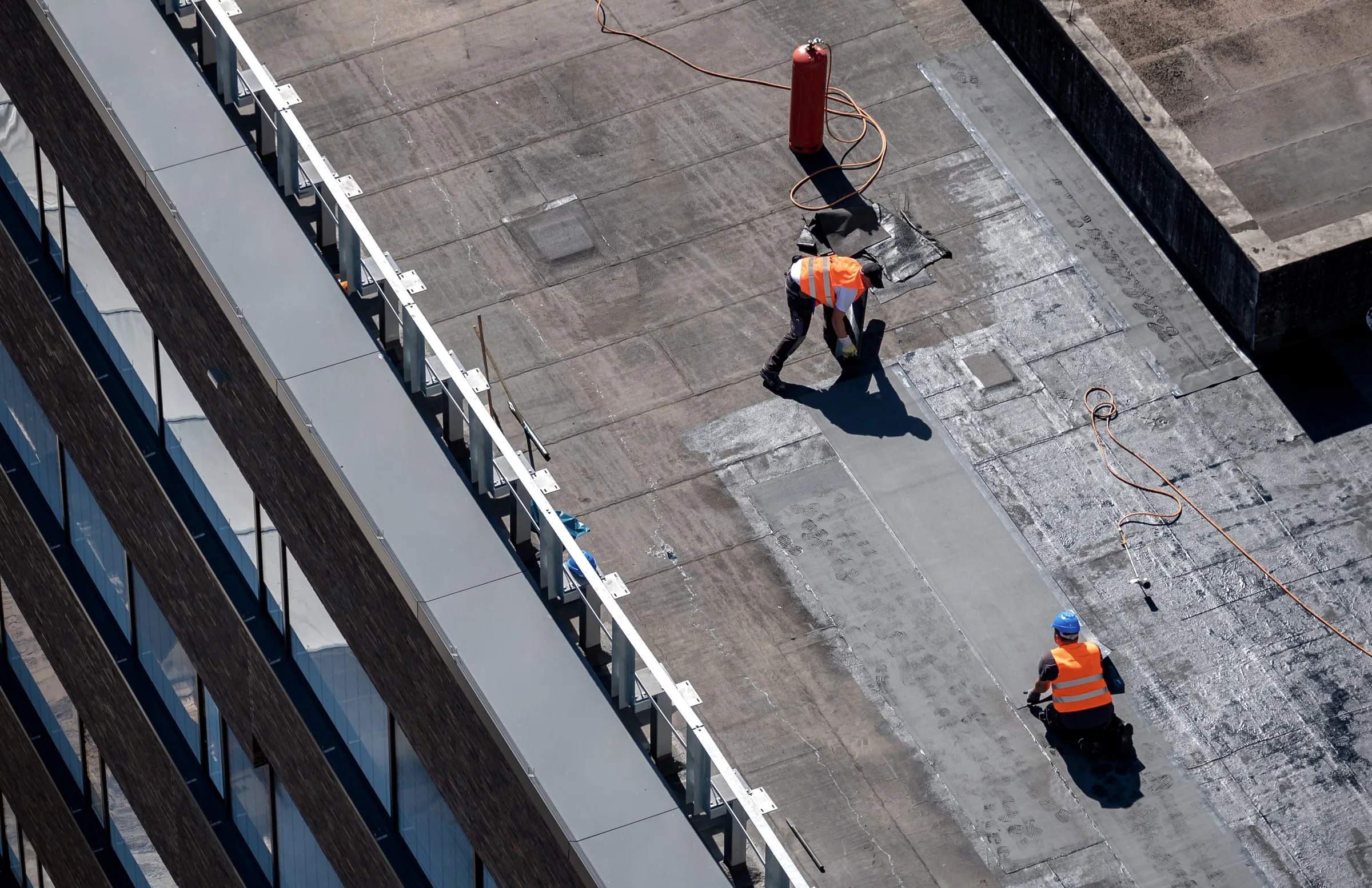 Aerial view of two roofers in orange safety vests working on a flat membrane roof beside a red torch tank and coiled hoses