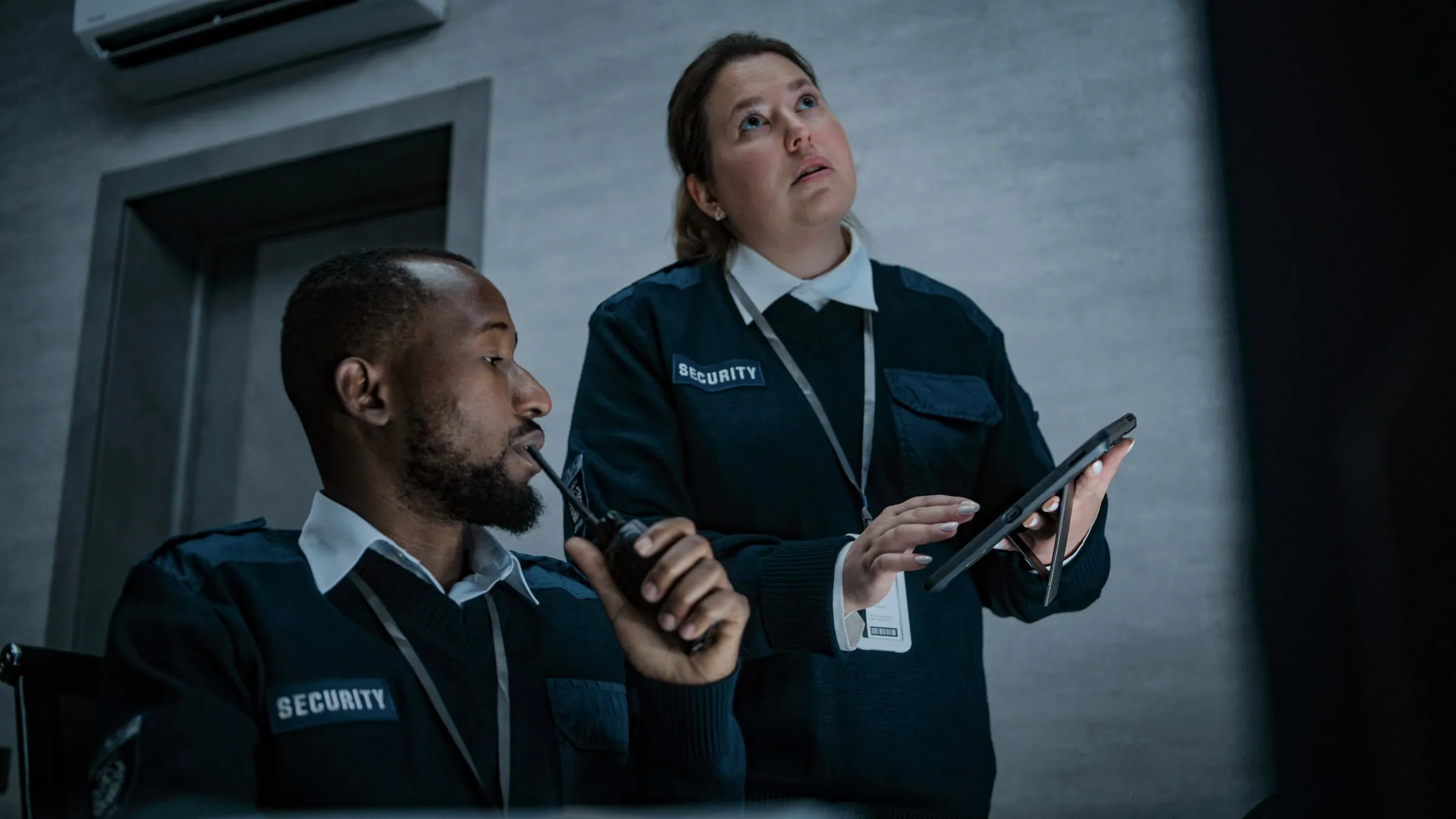 Two security officers in matching SECURITY uniforms, one speaking into a radio and one holding a tablet