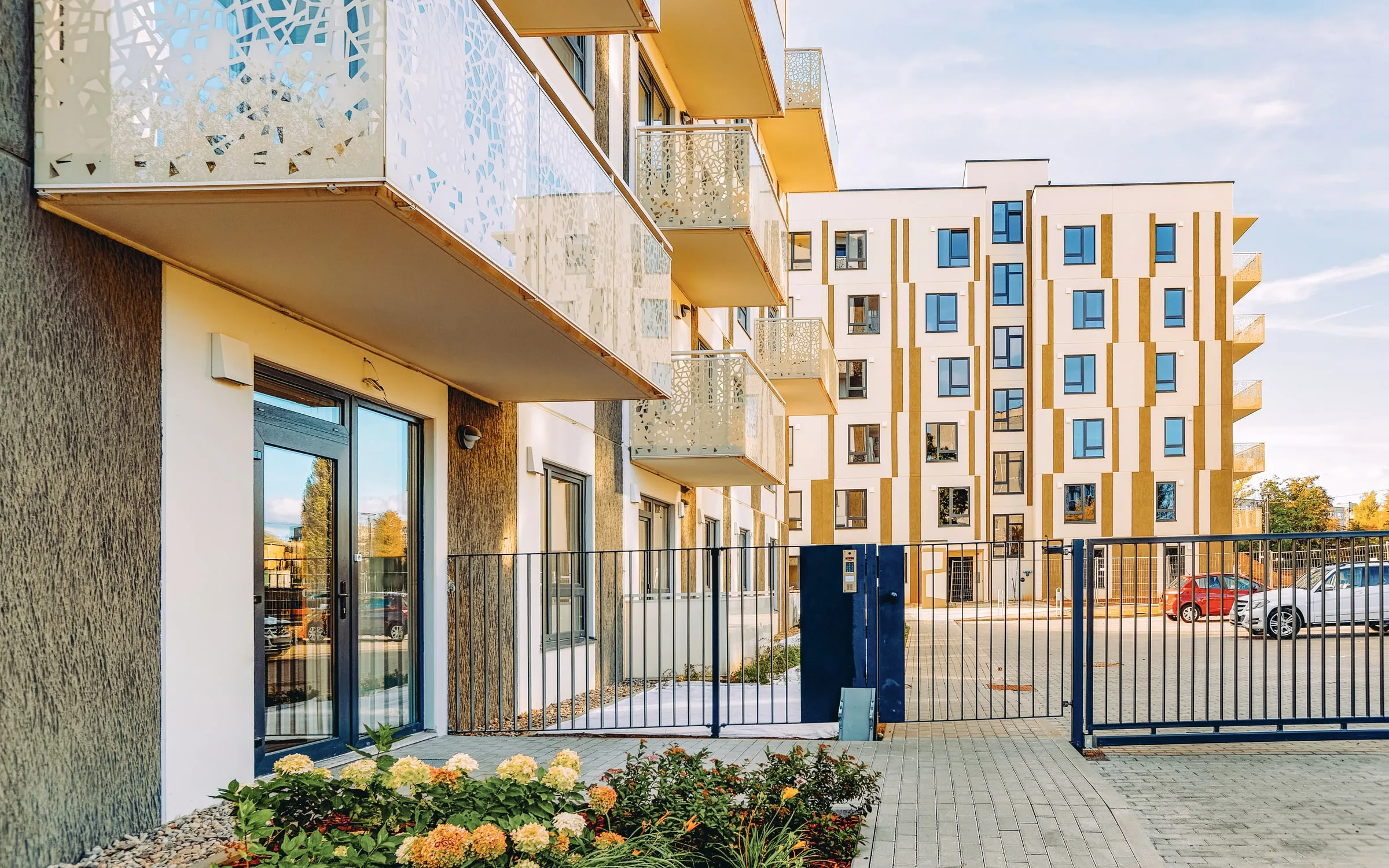 Mid-rise apartment buildings behind a metal fence and gate with a flower bed in the foreground