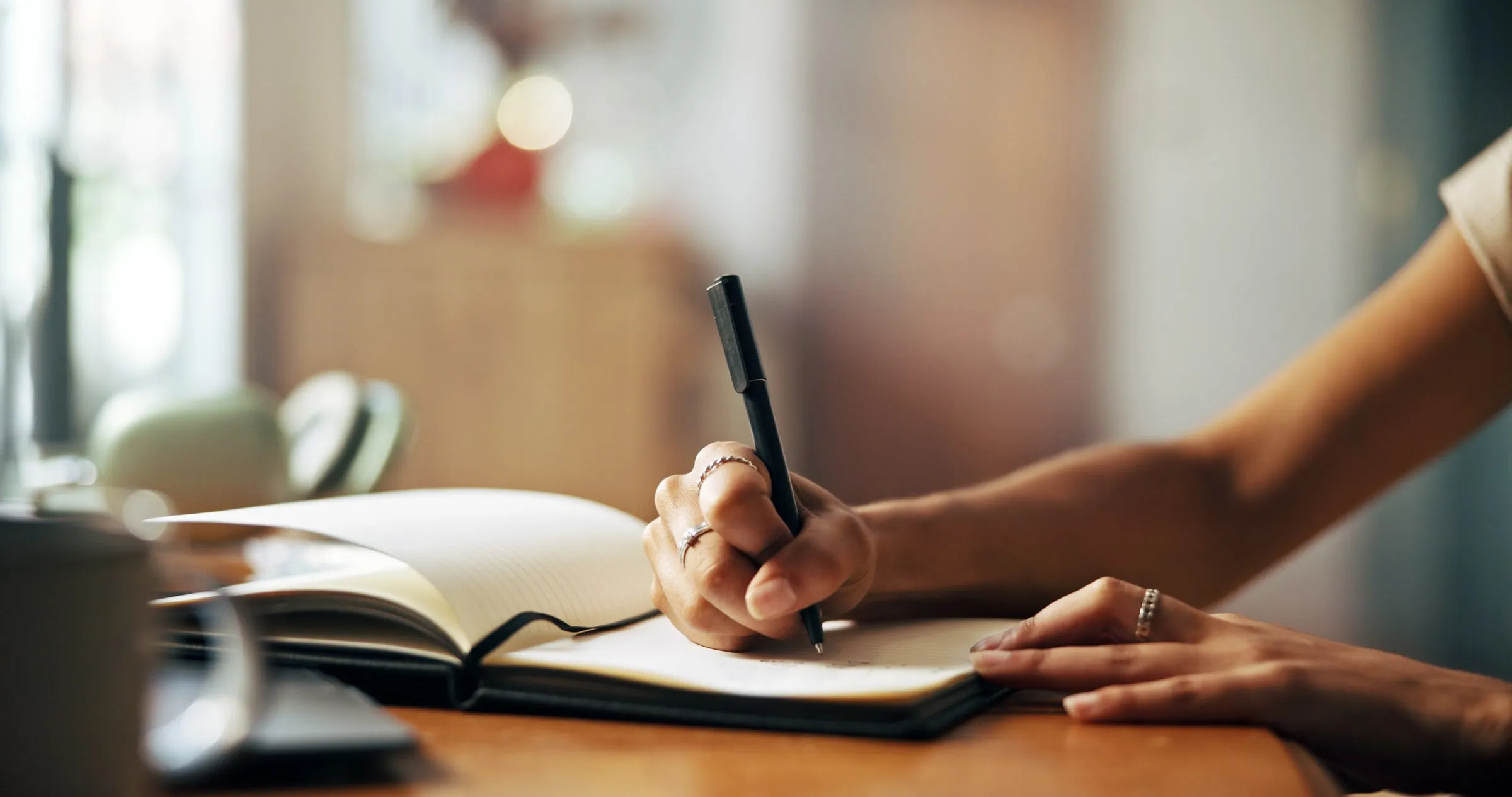 Person writing in a bound notebook with a pen at a wooden desk