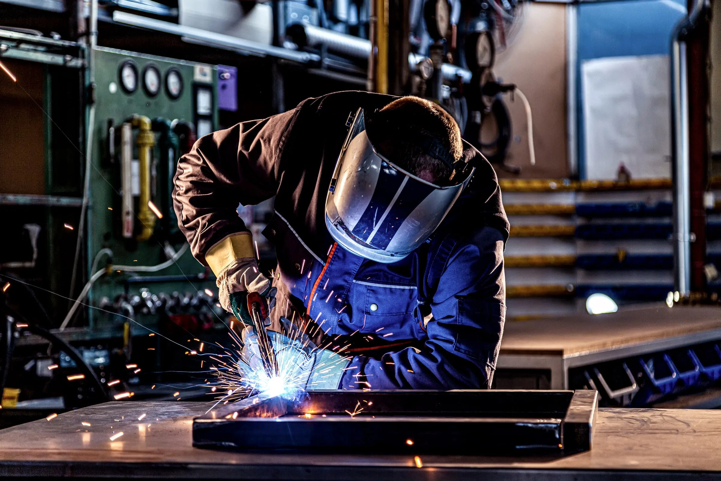 Welder in a hooded helmet and dark jacket welding a steel beam with bright sparks in a workshop
