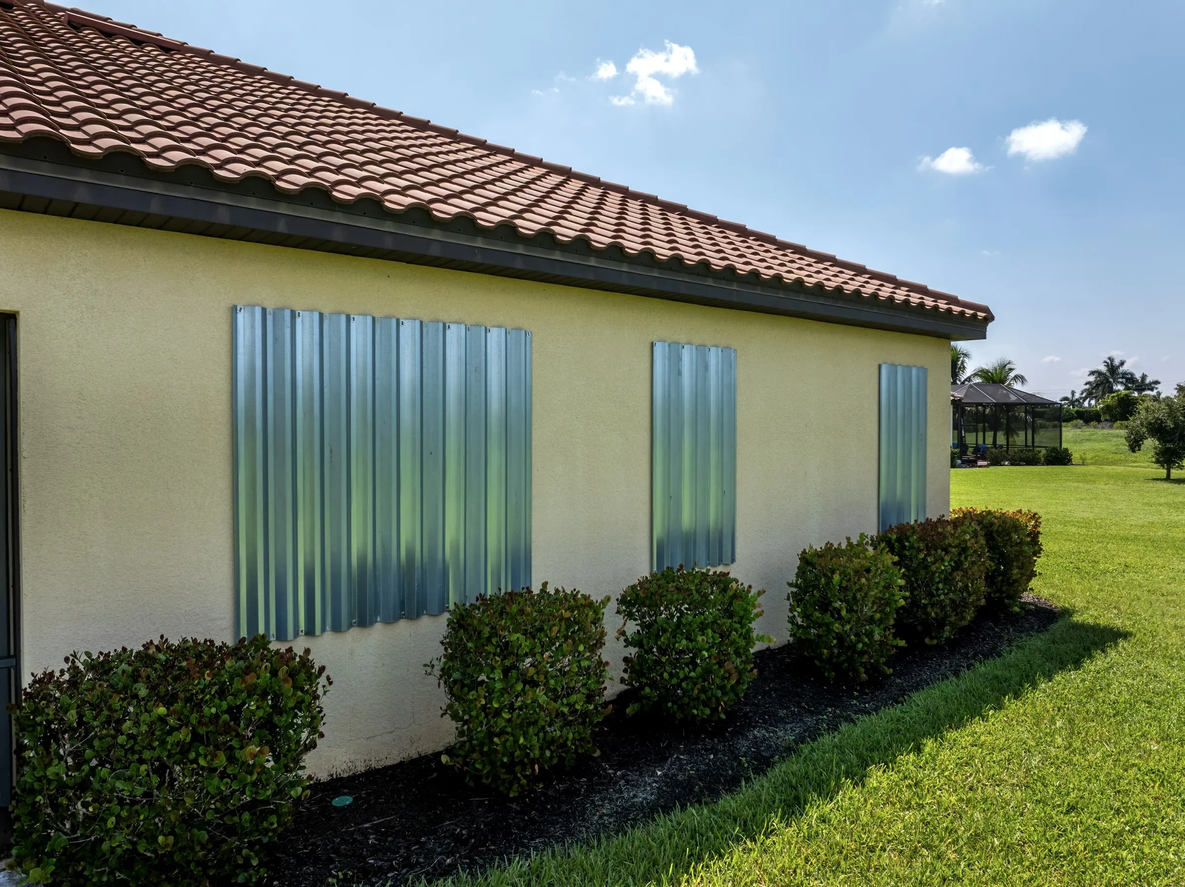Metal hurricane shutters covering the windows of a yellow house with a tiled roof