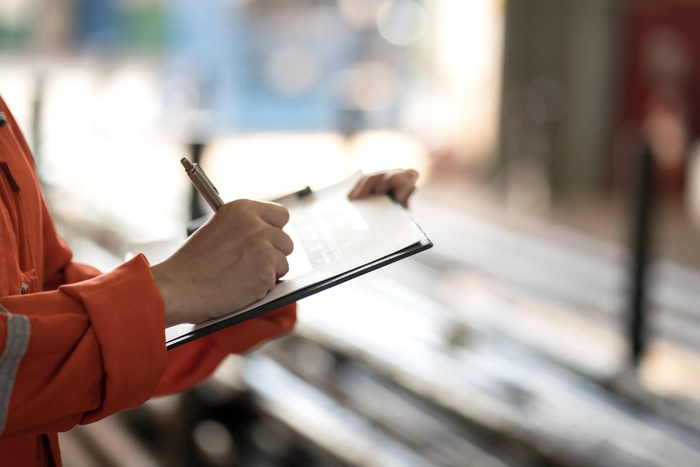 Person in an orange high-visibility jacket writing on a clipboard at an industrial site